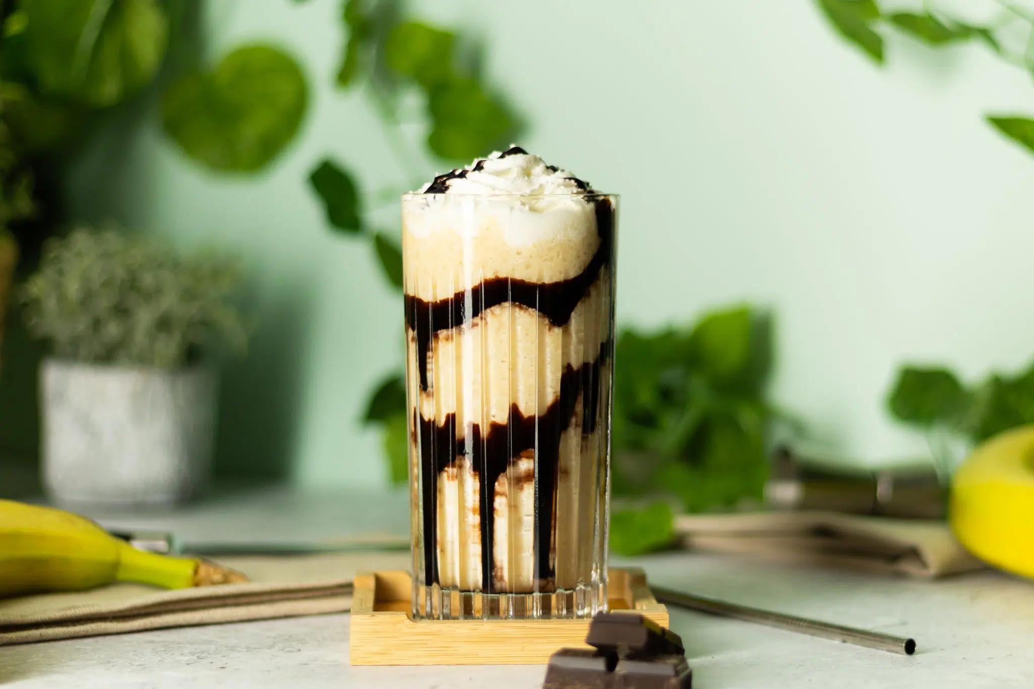 A side shot of a Dirty Banana cocktail on a coaster placed on a white table surrounded by chocolate squares, a straw, and two bananas, in front of a light green wall.