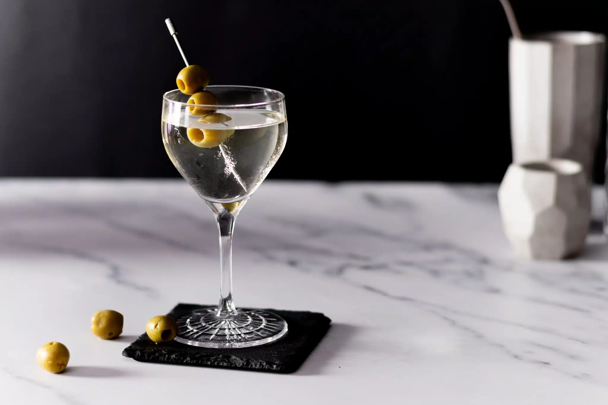 A side shot of a Dirty Martini in a coupe glass on a black stone coaster placed on a white marmol table with some olives on the side.