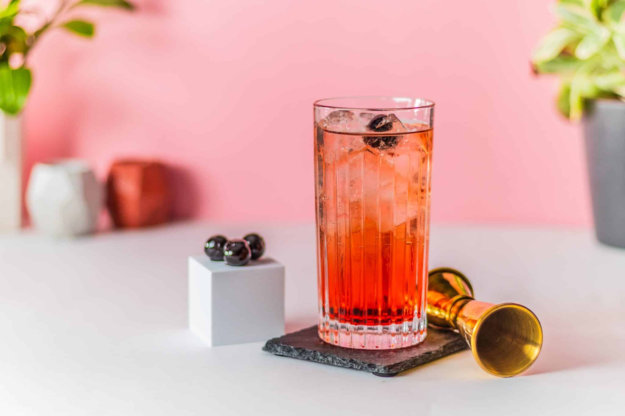 A side shot of a Dirty Shirley cocktail in a highball glass on a black stone plate with three cherries on a cube and a jigger on the side placed on a white table and a pink wall on the background.