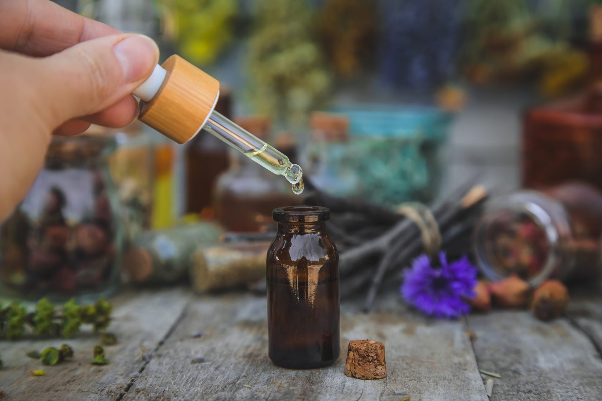 A shot of a hand holding a dropper and dropping some drops into the bottle surrounded by botanicals and herbals