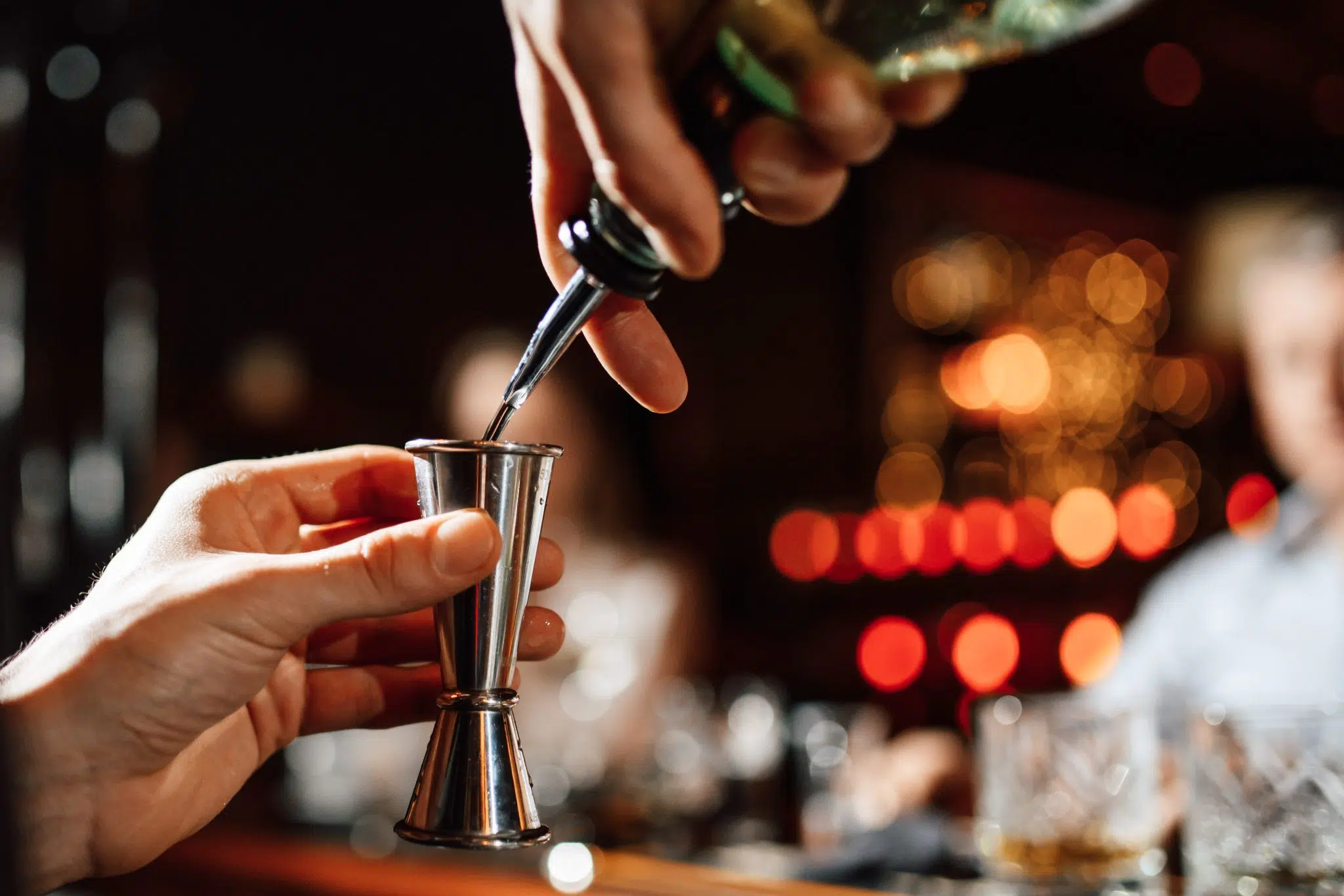 A shot of a bartender holding with one hand a double jigger and with the other hand a bottle with a spout pouring the liquid into the double jigger