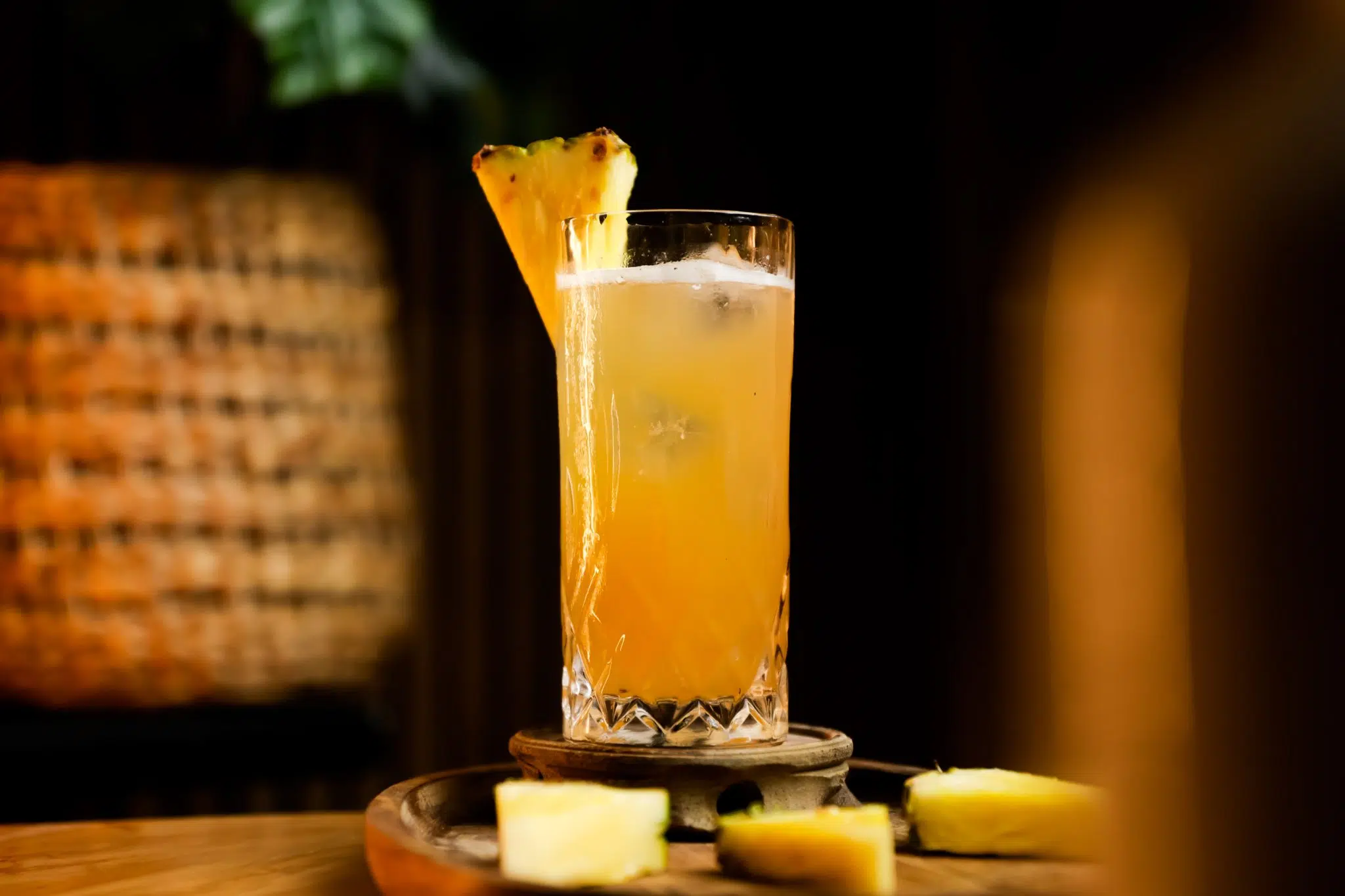 A side shot of a Drunken Monkey Rum cocktail in a highball glass on a wooden coaster surrounded by pineapple wedges
