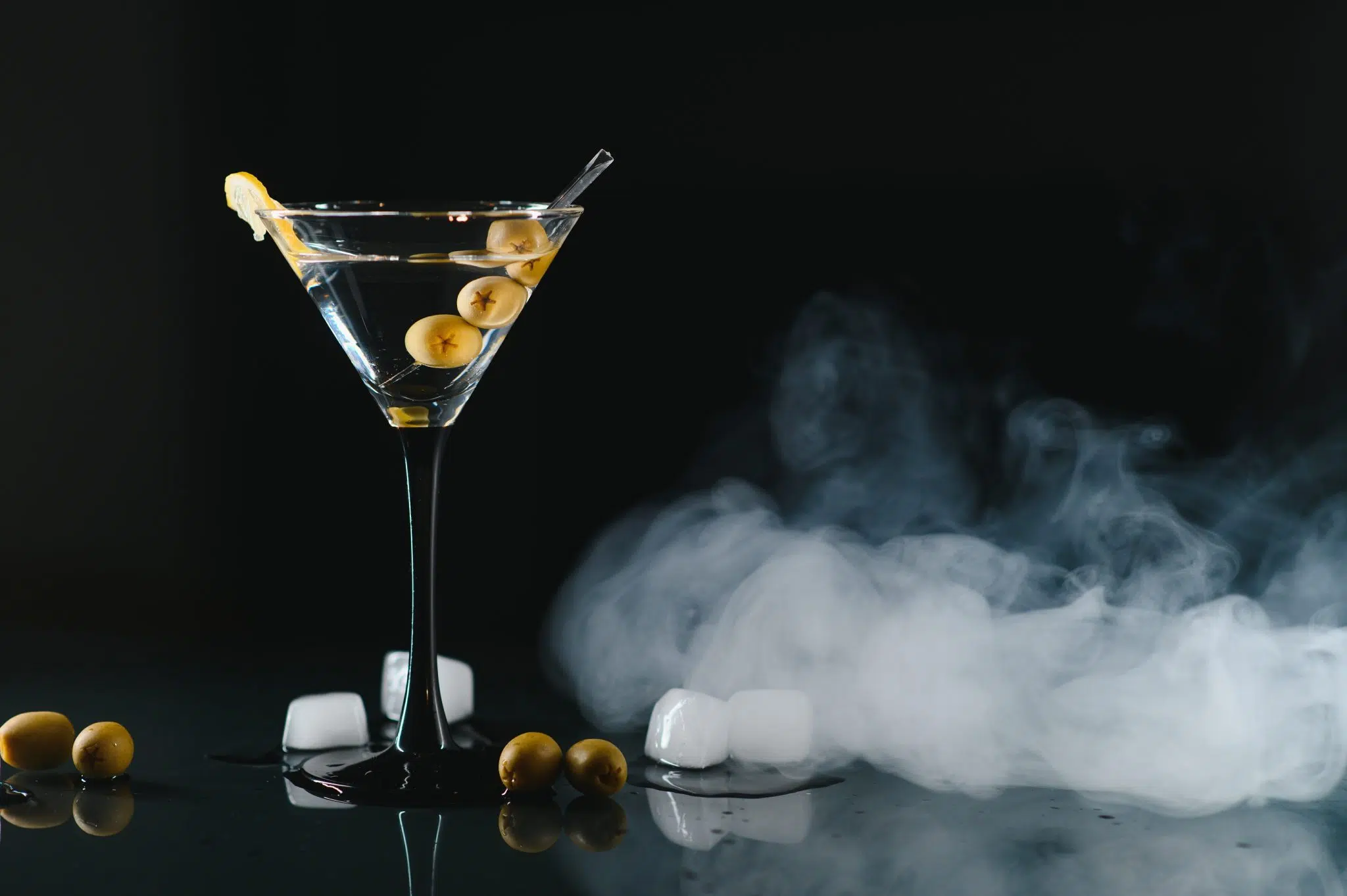 A side shot of a Dry Ice Martini cocktail in a martini glass surrounded by olives and dri ice cubes on a black background
