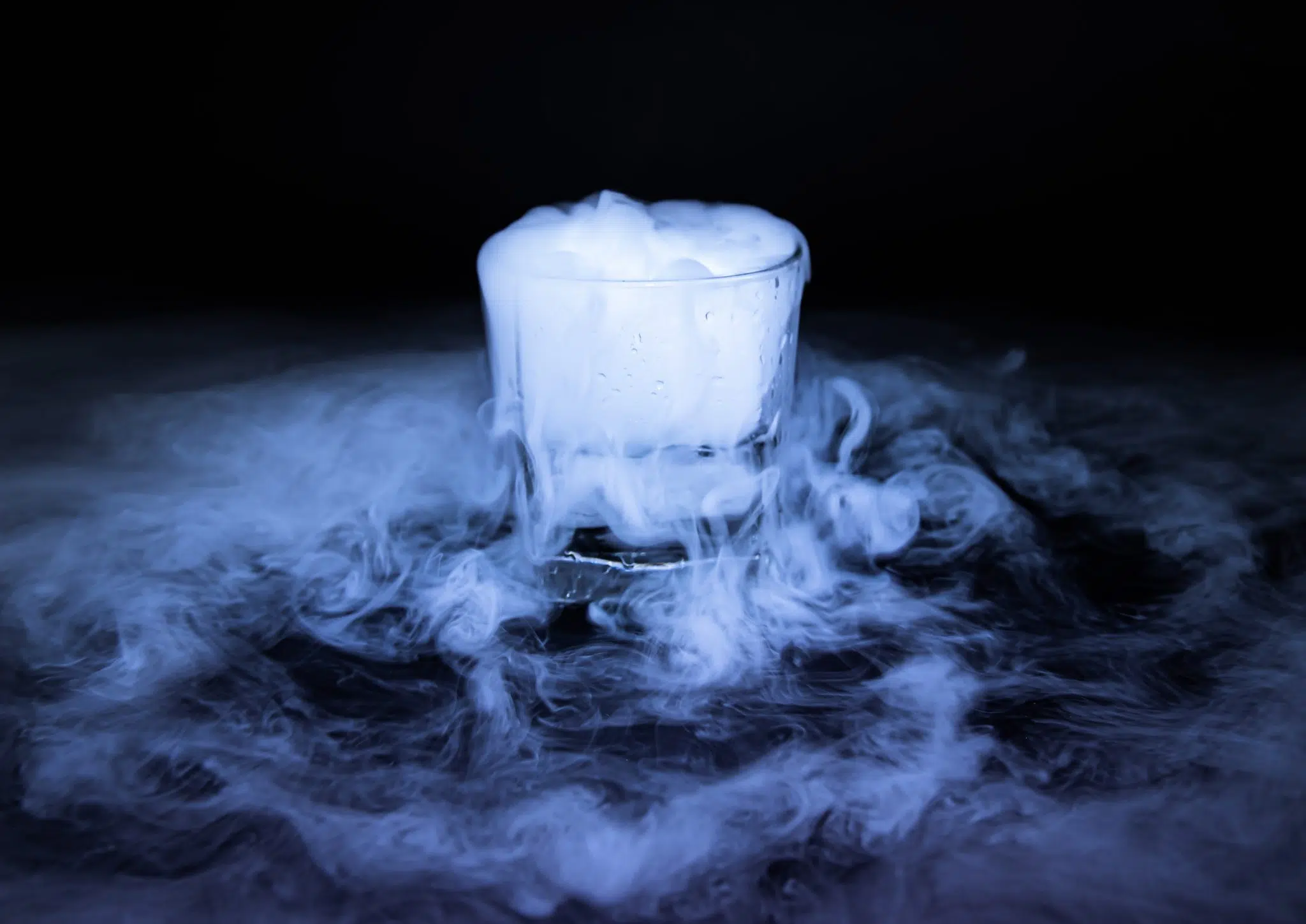 A side shot of a glass chilled with a dry ice cube with a smoke effect, placed on a black surface and background