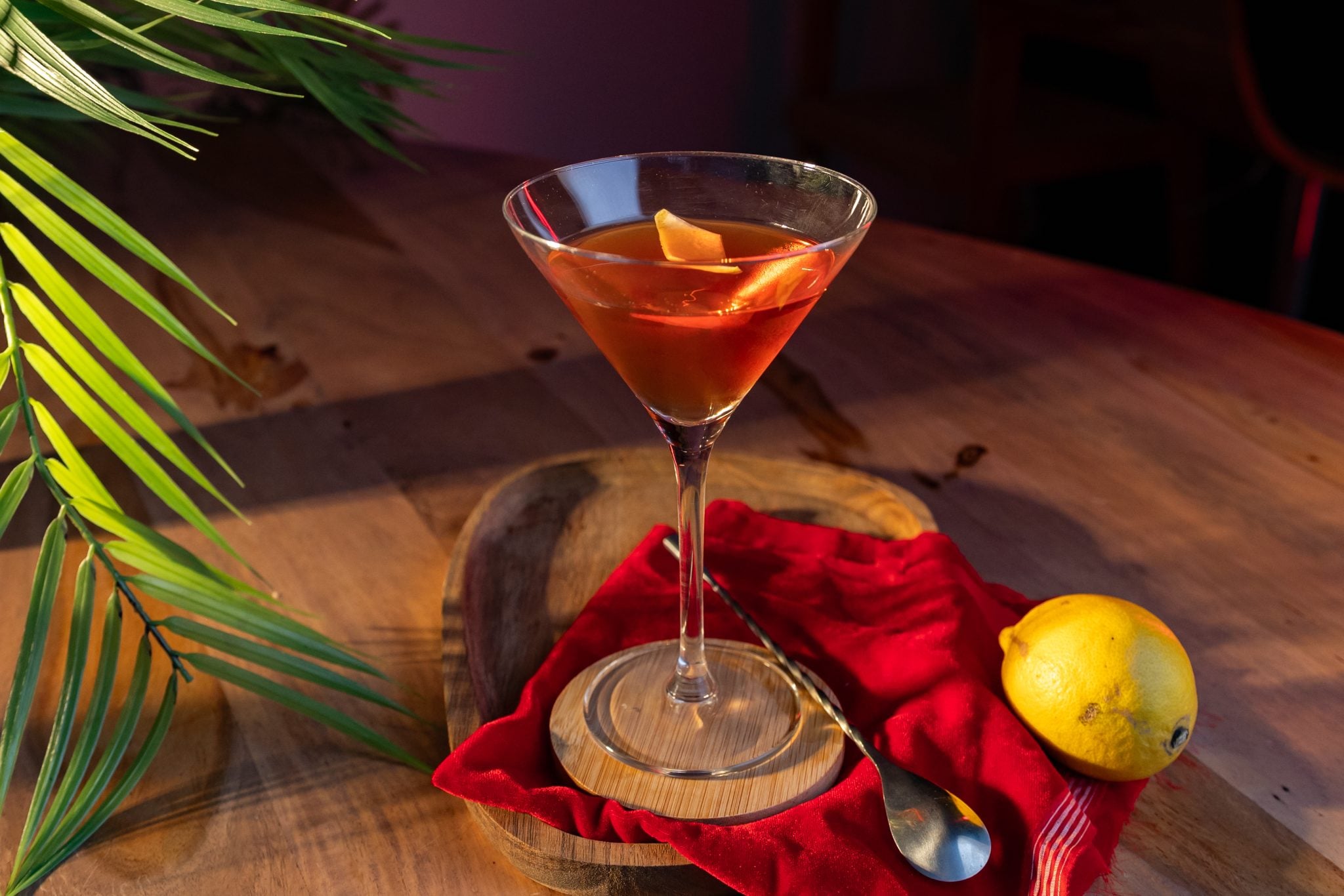 A side shot of a Dry Rob Roy cocktail in a martini glass on a wooden coaster placed on a wooden table surrounded by a red cloth, a lemon, and a bar spoon