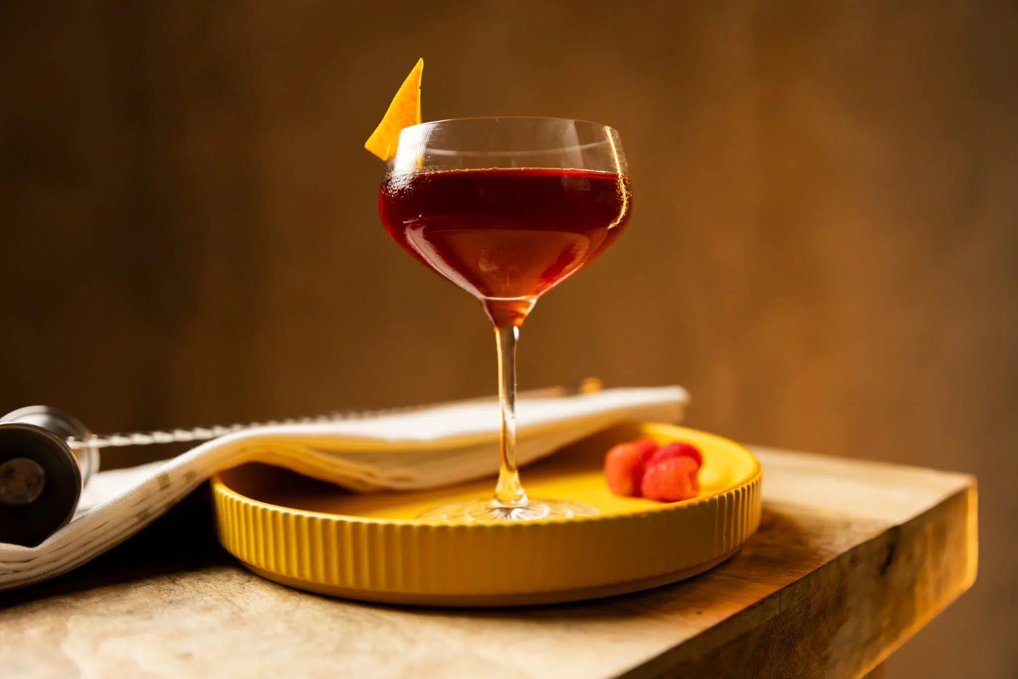 A side shot of an East India No.1 cocktail in a coupe glass on a yellow tray surrounded by three raspberries, a white cloth and a jigger