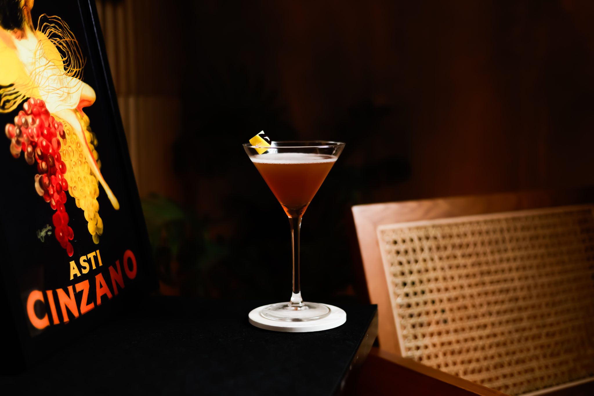 A side shot of a East India No.2 cocktail in a martini glass on a white coaster placed on a black surface with a frame and a chair around