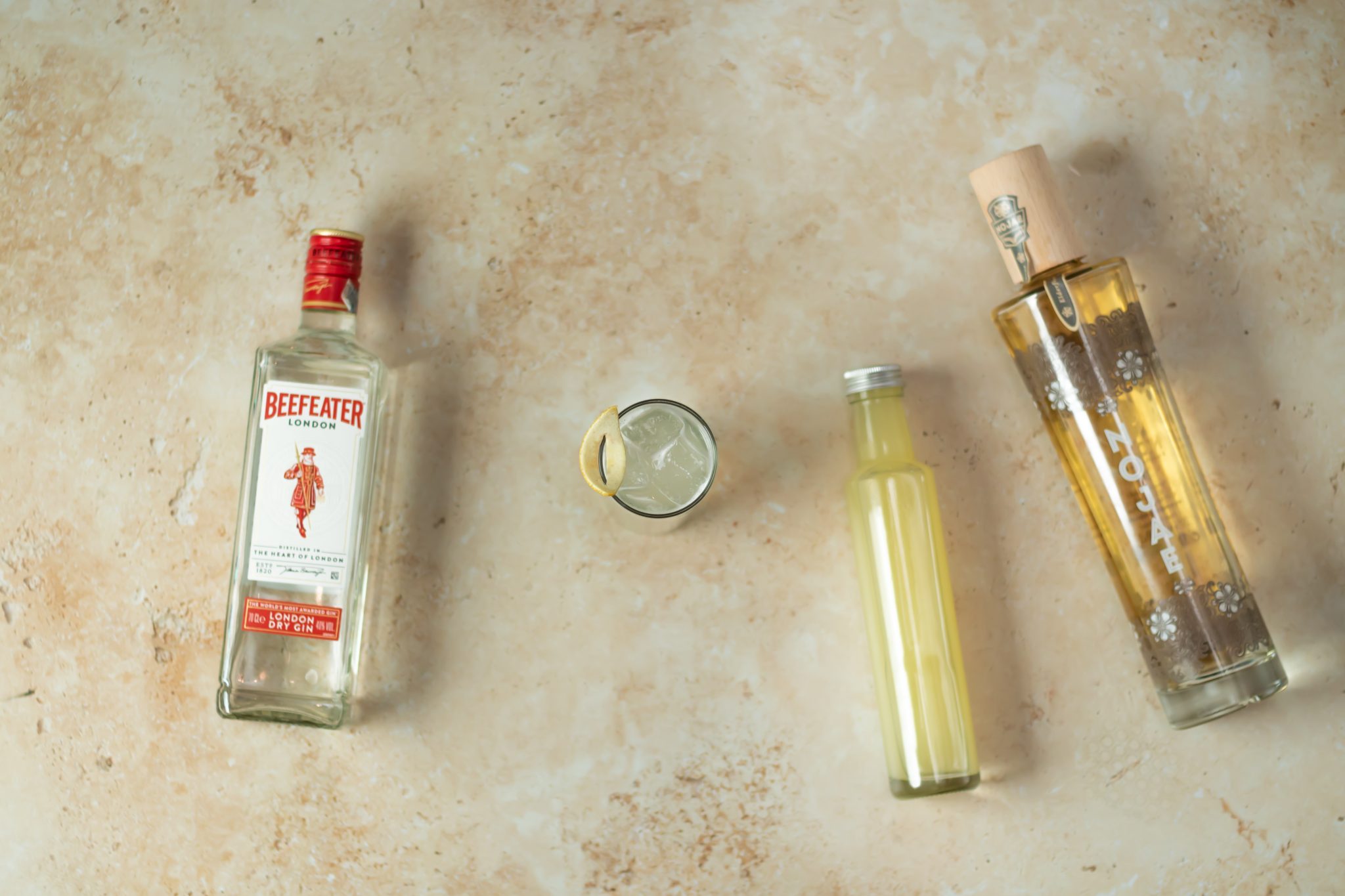 An Elderflower Collins cocktail, shot from above, in a highball glass on a beige surface surrounded by Gin, lemon juice, and elderflower liqueur.