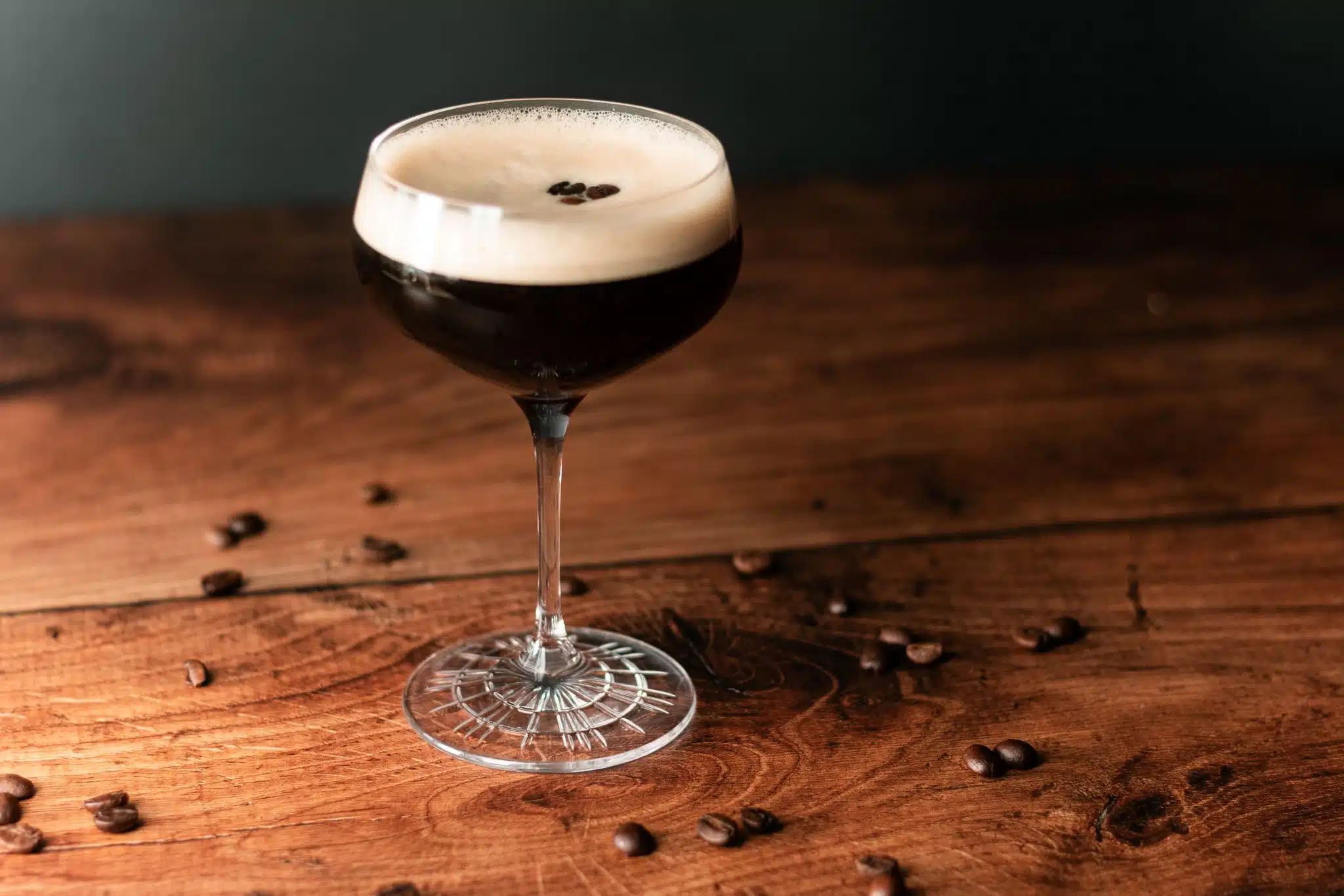 A side shot of a Espresso Martini cocktail in a martini glass on a dark wooden table surrounded by coffee beans.
