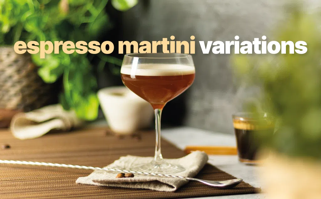 An espresso martini cocktail in a cocktail glass on a beige cloth placed on a brown placemat surrounded by an espresso, a bar spoon, and two coffee beans
