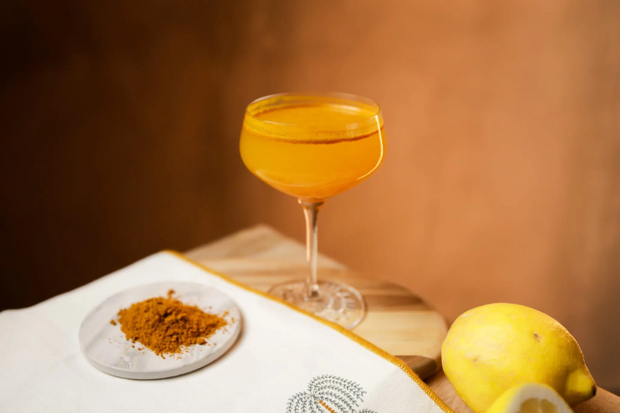 A side shot of a Everest cocktail in a coupe glass on a wooden board and table surrounded by a lemona white cloth and curry powder on a white coaster