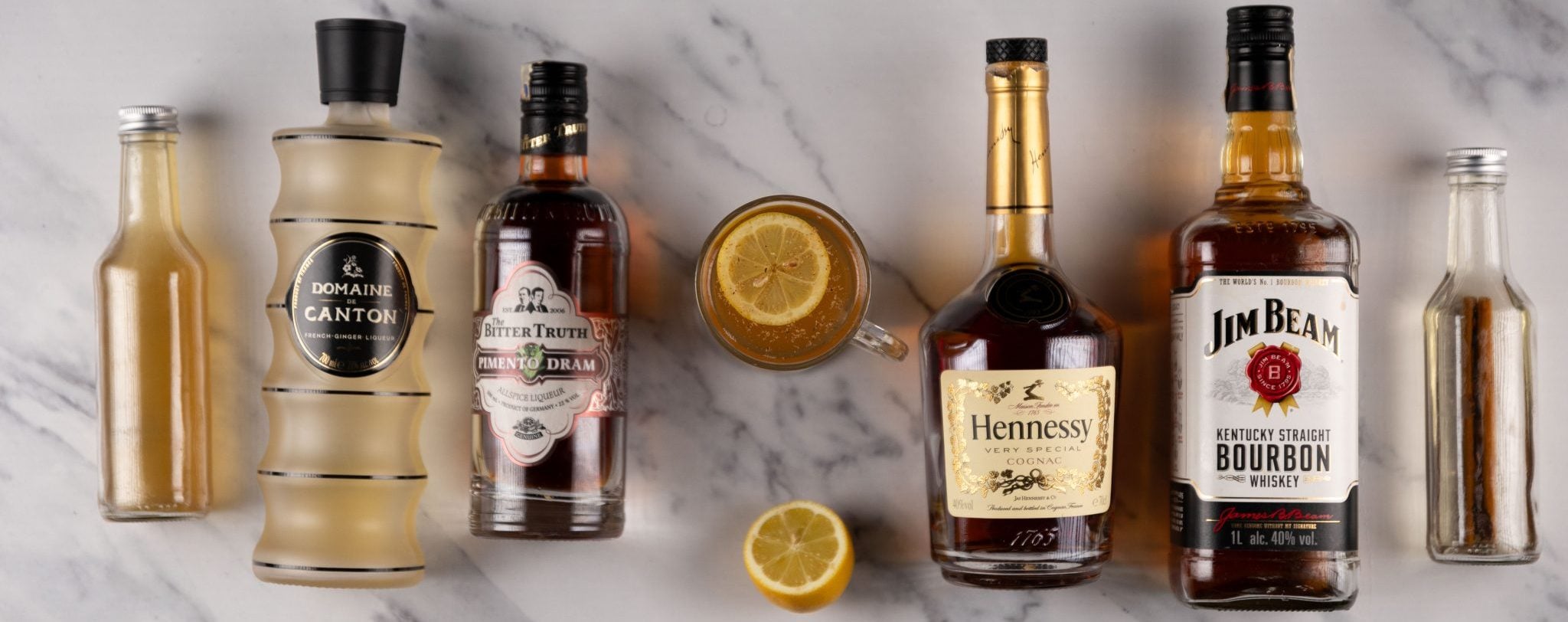 A Fallen Leaf cocktail, shot from above, in a mug on a white marmol surface surrounded by Bourbon, brandy de Jerez, ginger liqueur, allspice dram liqueur, lemon, honey syrup, cinnamon lemon infused hot water