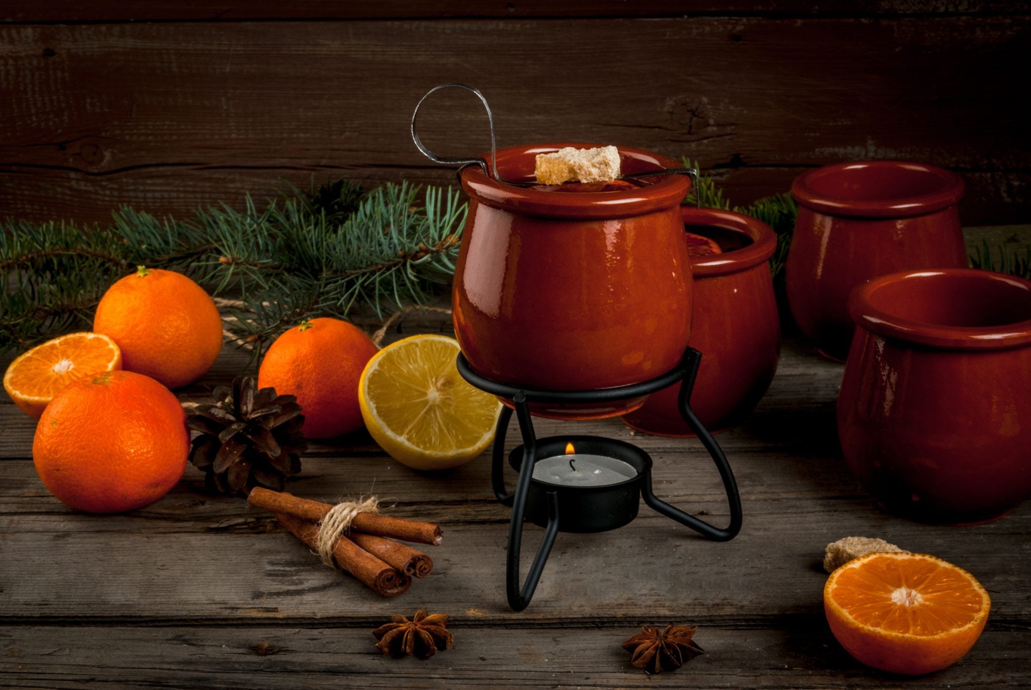A clay mug with Feuerzangenbowle with a candle below with oranges, lemons, anise stars and cinnamon sticks