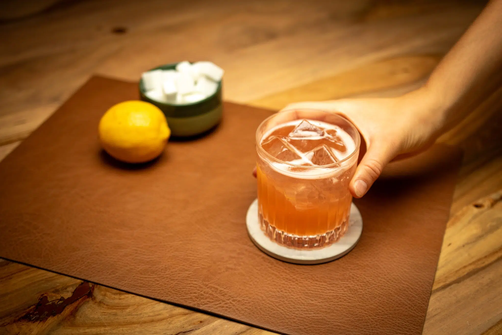 A side shot of a Fitzgerald cocktail in an Old Fashioned glass holded by a hand on a white coaster placed on a brown placemat on a wooden table with a lemon and a green bolw with sugar cubes inside