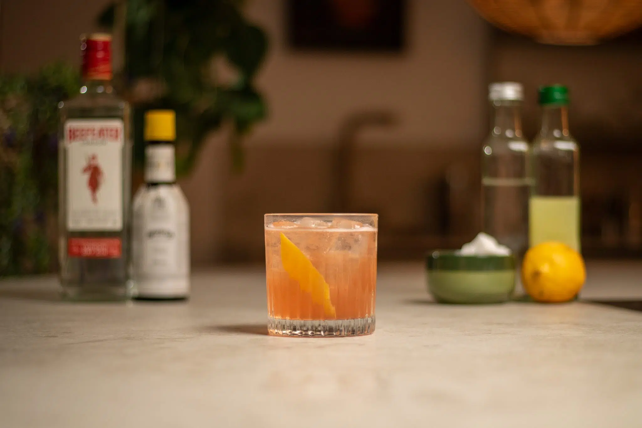 Gin, lemon juice, lemon, simple syrup, sugar cubes in a bowl, and Angostura bitters laid out on a white bar table