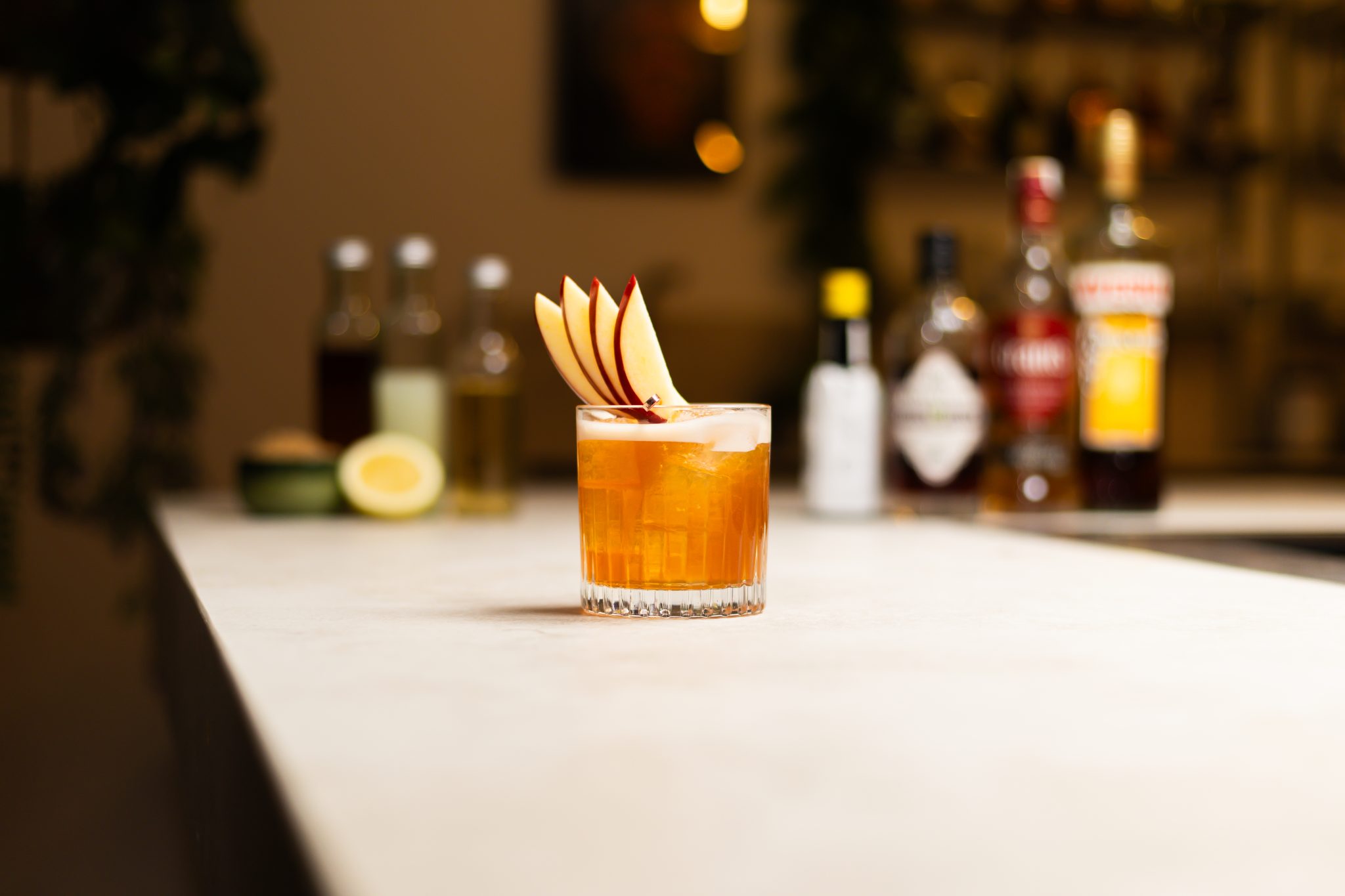 Scotch whisky, apple cider, lemon juice, lemon, Demerara sugar syrup, allspice dram, Averna Amaro, and Angostura bitters laid out on a white bar table