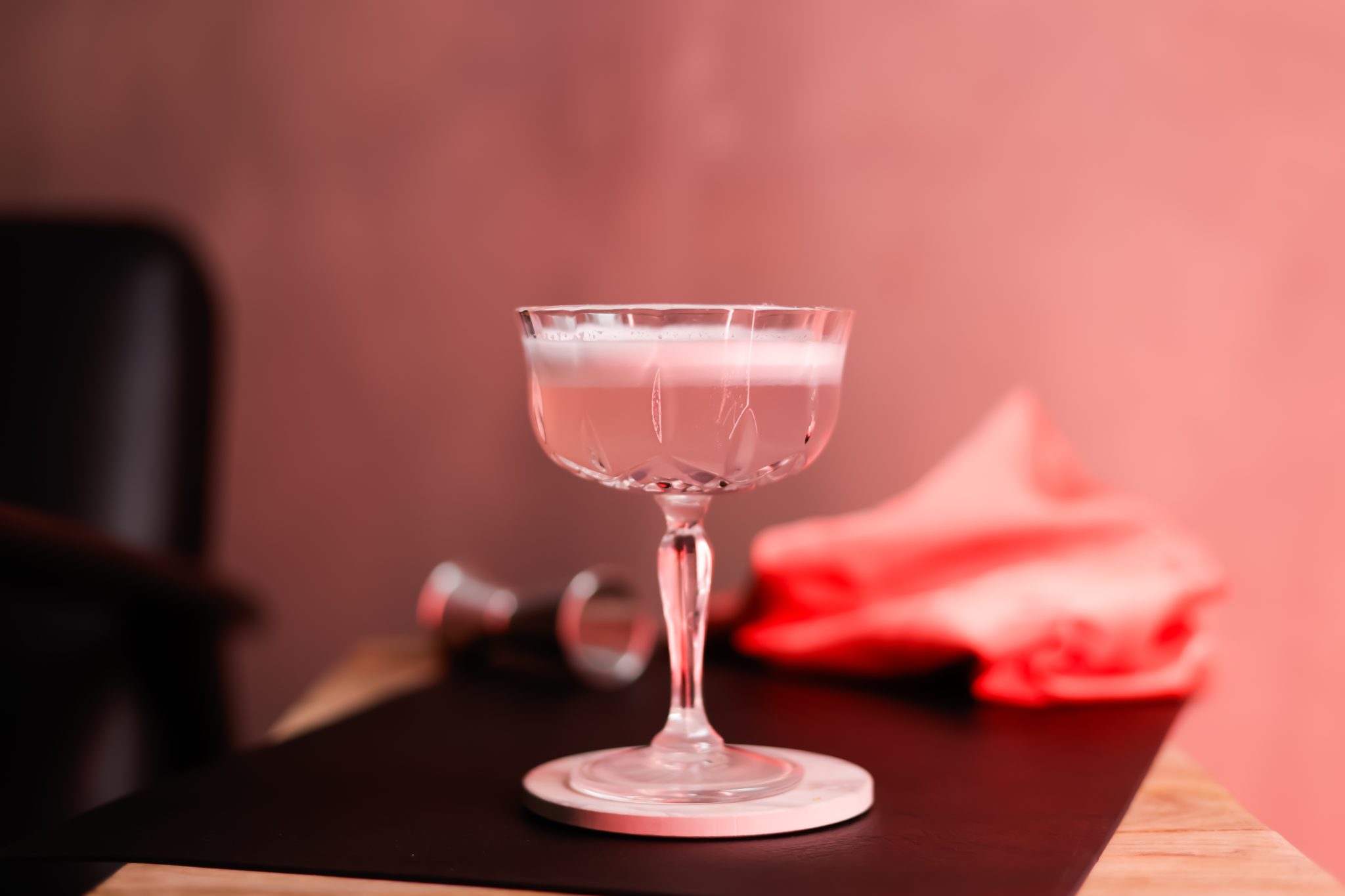 A side shot of a Flower Power Sour cocktail in a coupe glass on a brown placemat surrounded by a jigger and a pink cloth, in front of a pink background