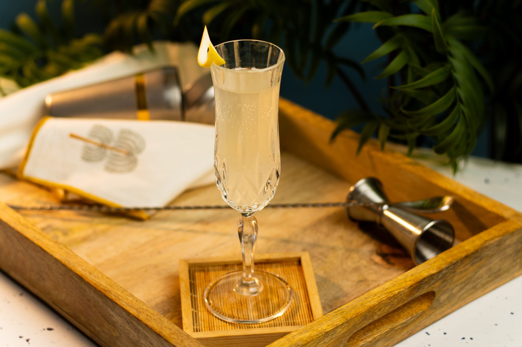 A side shot of a French 75 cocktail in a champagne flute on a wooden coaster placed on a wooden tray surrounded by a jigger, a bar spoon and a white cloth