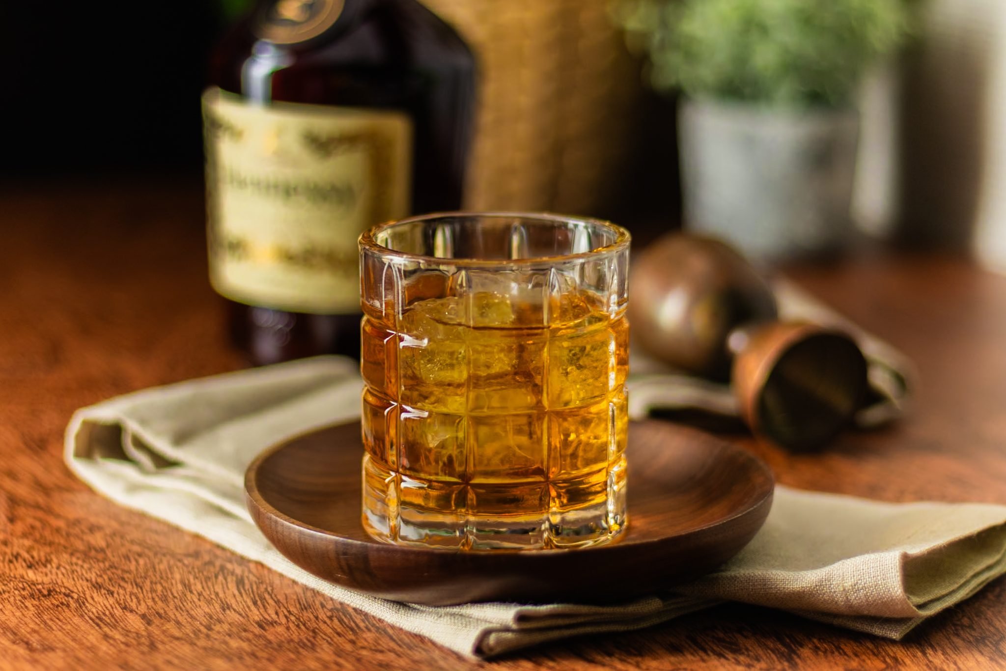 A side shot of a French Connection cocktail in an Old Fashioned glass on a plate placed on a white cloth on a wooden table with a Hennessy Cognac bottle and a jigger around.