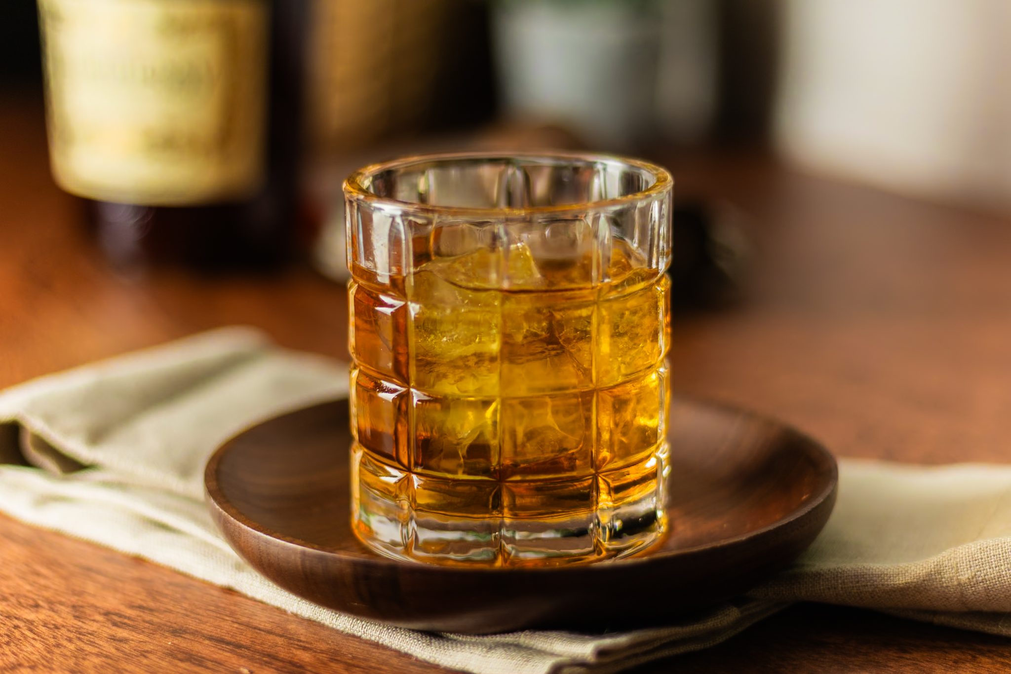 A side shot of a French Connection cocktail in an Old Fashioned glass on a plate placed on a white cloth on a wooden table with a Hennessy Cognac bottle around.