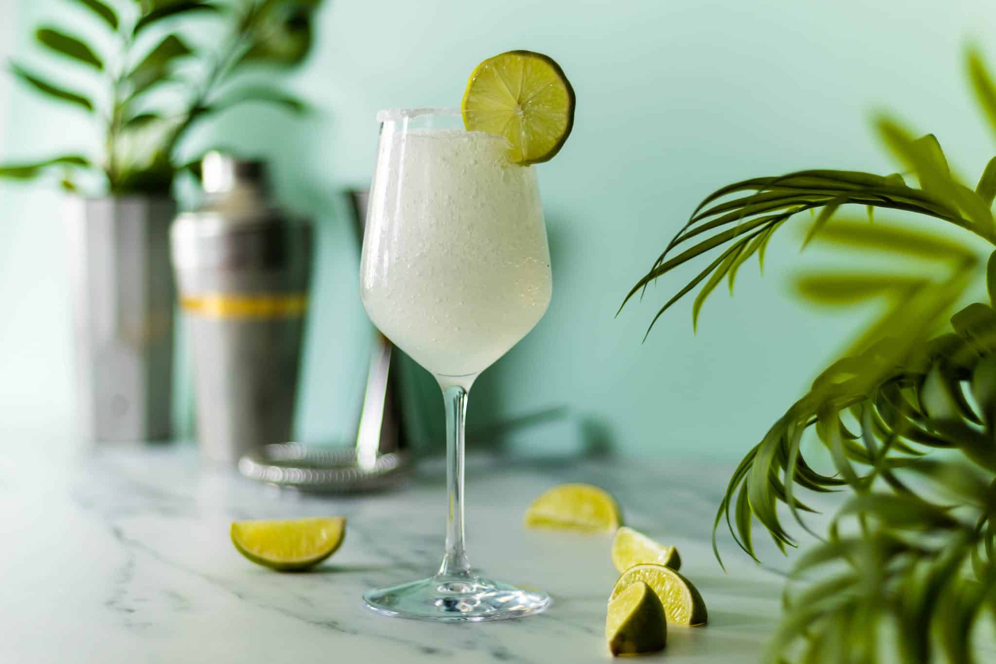 A side shot of a Frozen Margarita cocktail in a margarita glass on a white marmol table surrounded by lime wedges and a shaker on the background.