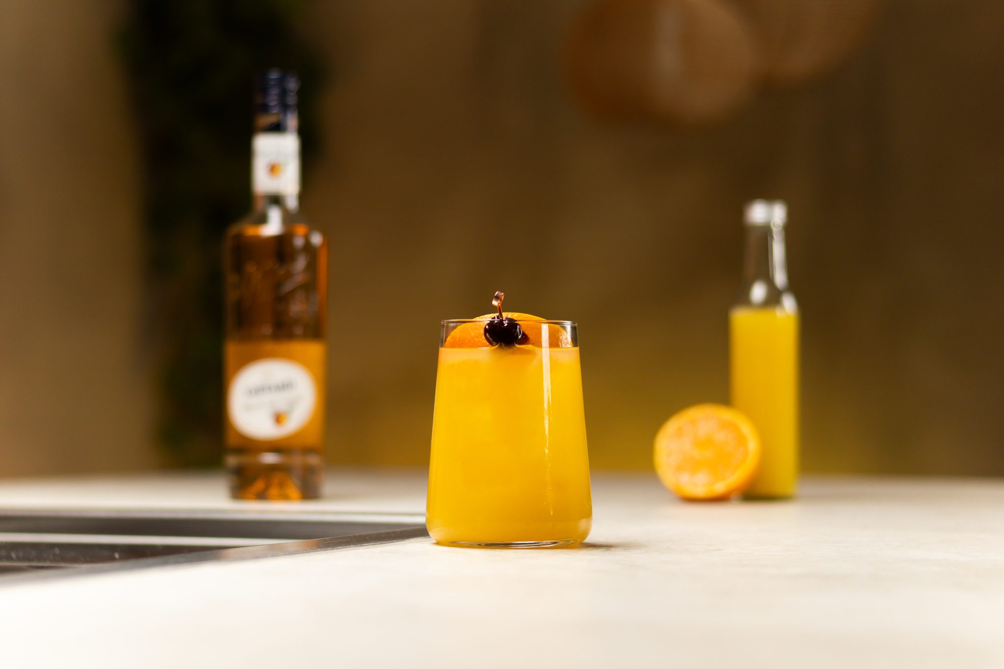 Peach Schnapps, orange juice and an orange laid out on a white bar table