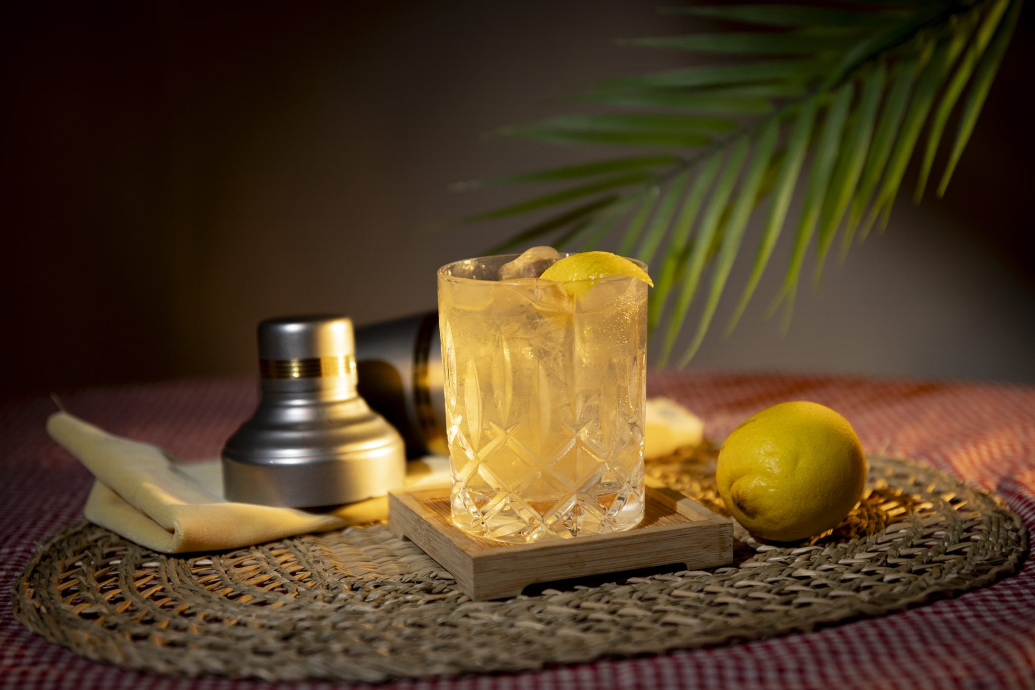 A side shot of a Garden Sour cocktail in an old fashioned glass on a wooden coaster placed on a straw placemat surrounded by a lemon, a shaker and a yellow cloth