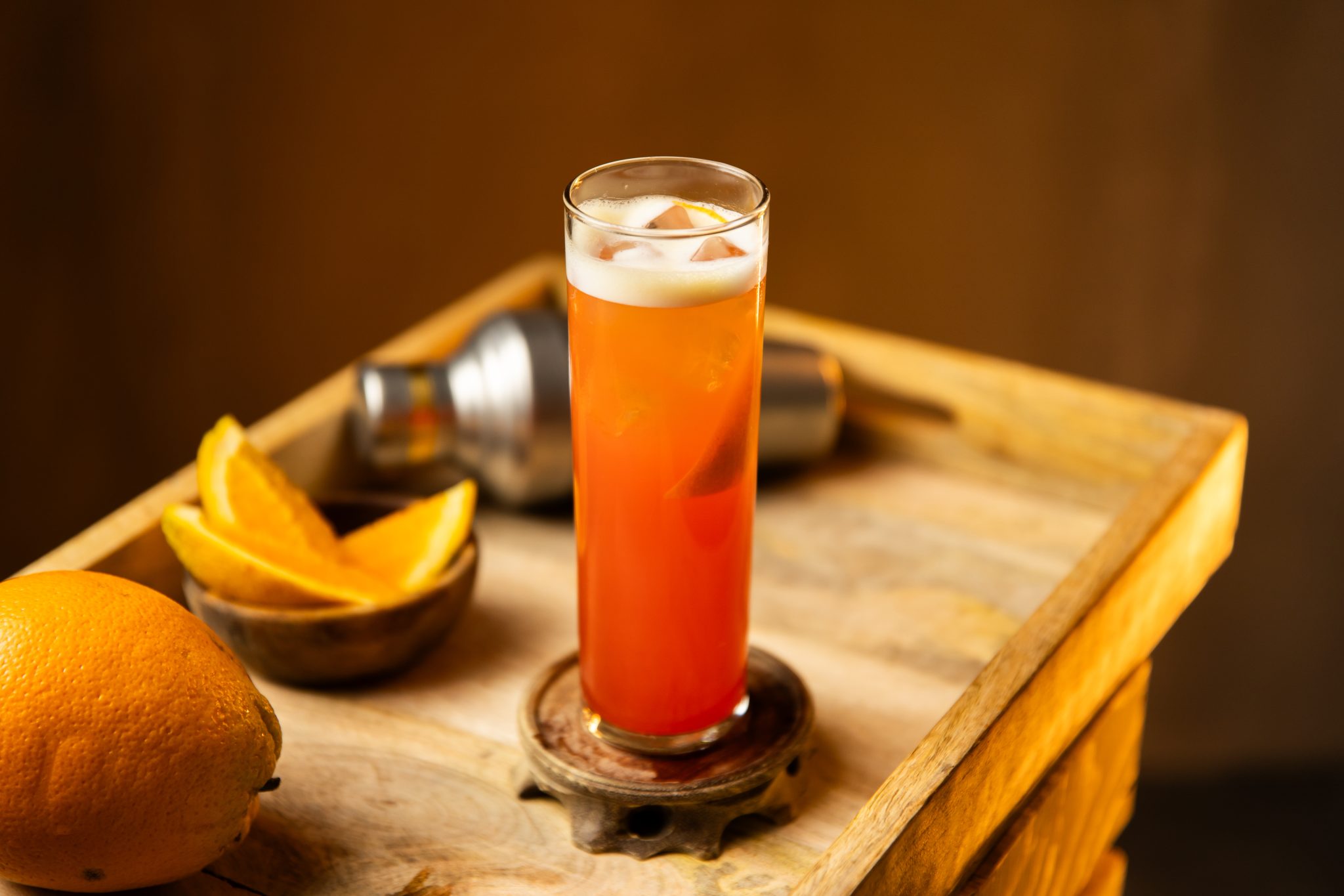 A side shot of a Garibaldi cocktail in a highball glass on a turquoise placemat placed on a table surrounded by a jigger, a bar spoon and a green bowl with orange wedges