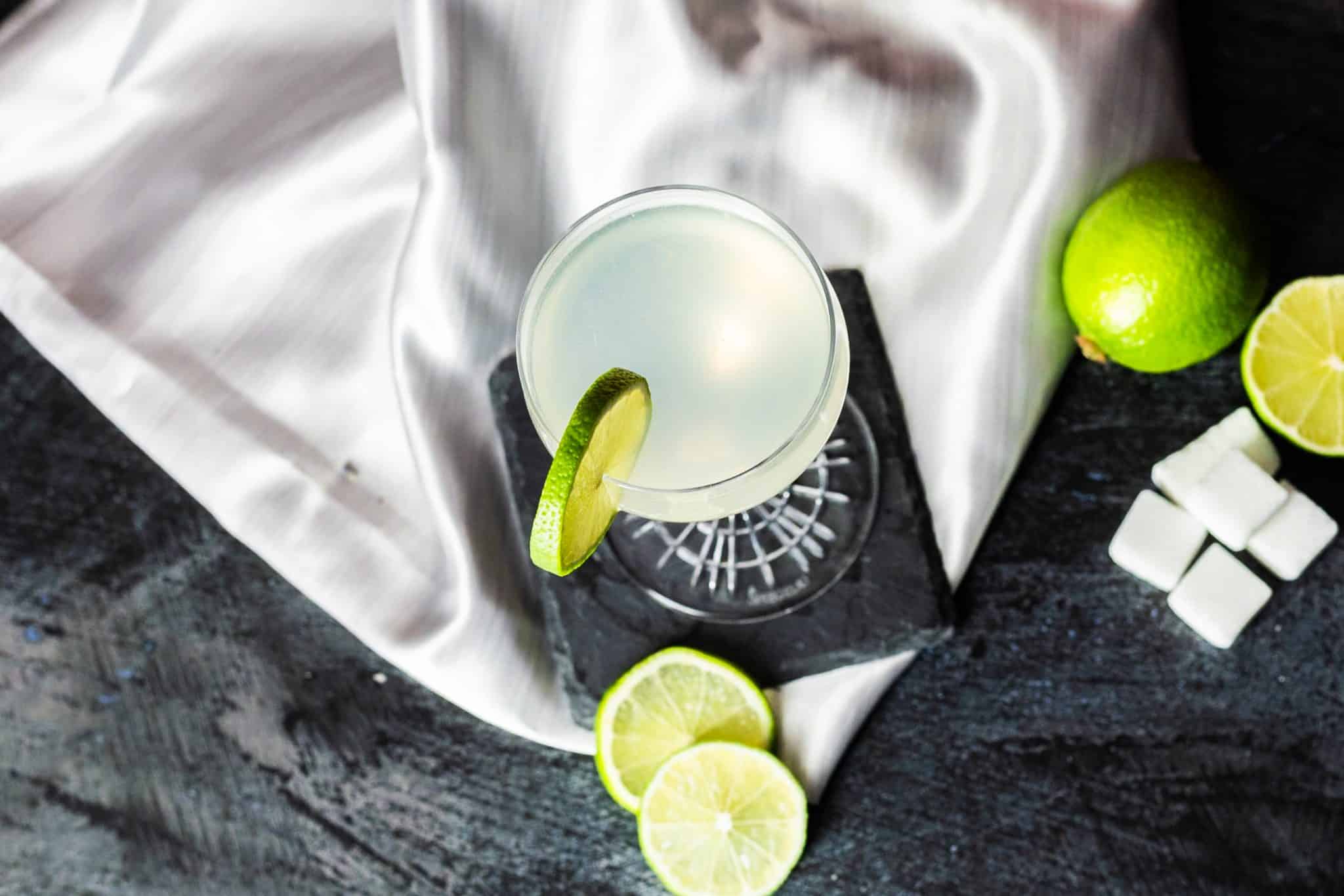 A Gimlet cocktail, shot from above, in a coupe glass on a black stone coaster placed on a black table with lime pieces, five sugar cubes, and a white cloth around.