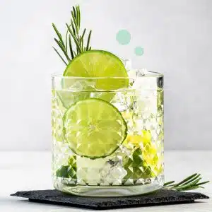 Old-fashioned glass filled with two limes wedges, ice cubes and gin spirit on a black stone coaster on a white table and in front of a white wall.