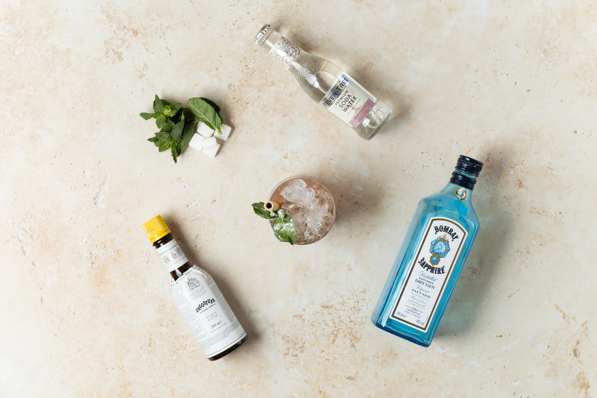 A Gin Julep cocktail, shot from above, in a old fashioned glass on a beige surface surrounded by London Dry Gin, mint leaves, sugar cube, soda water and Angostura aromatic bitters