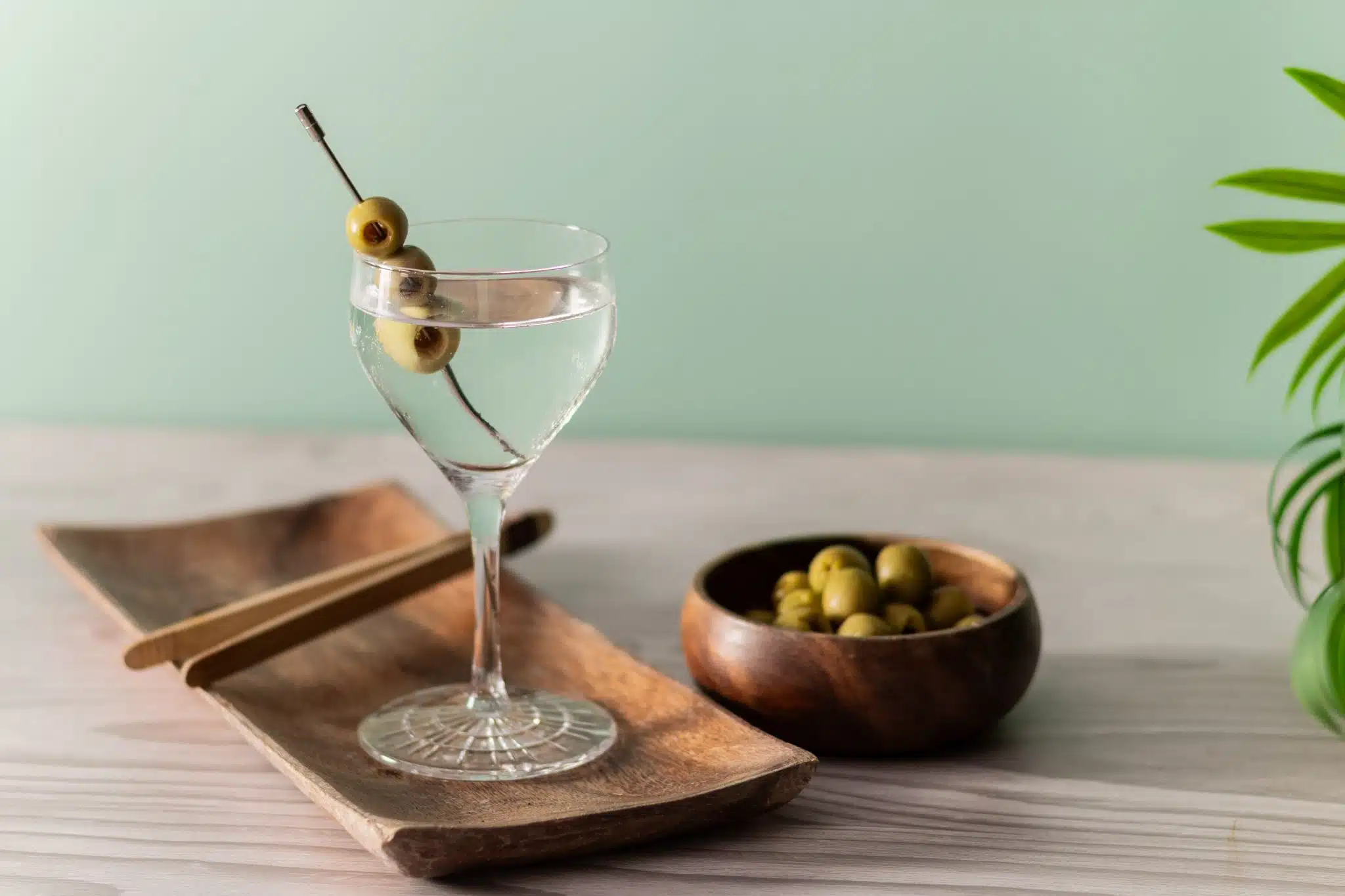 A side shot of a Gin Martini cocktail in a martini glass on a wooden tray with a bowl with olives placed on a table and a turquoise wall on the background.