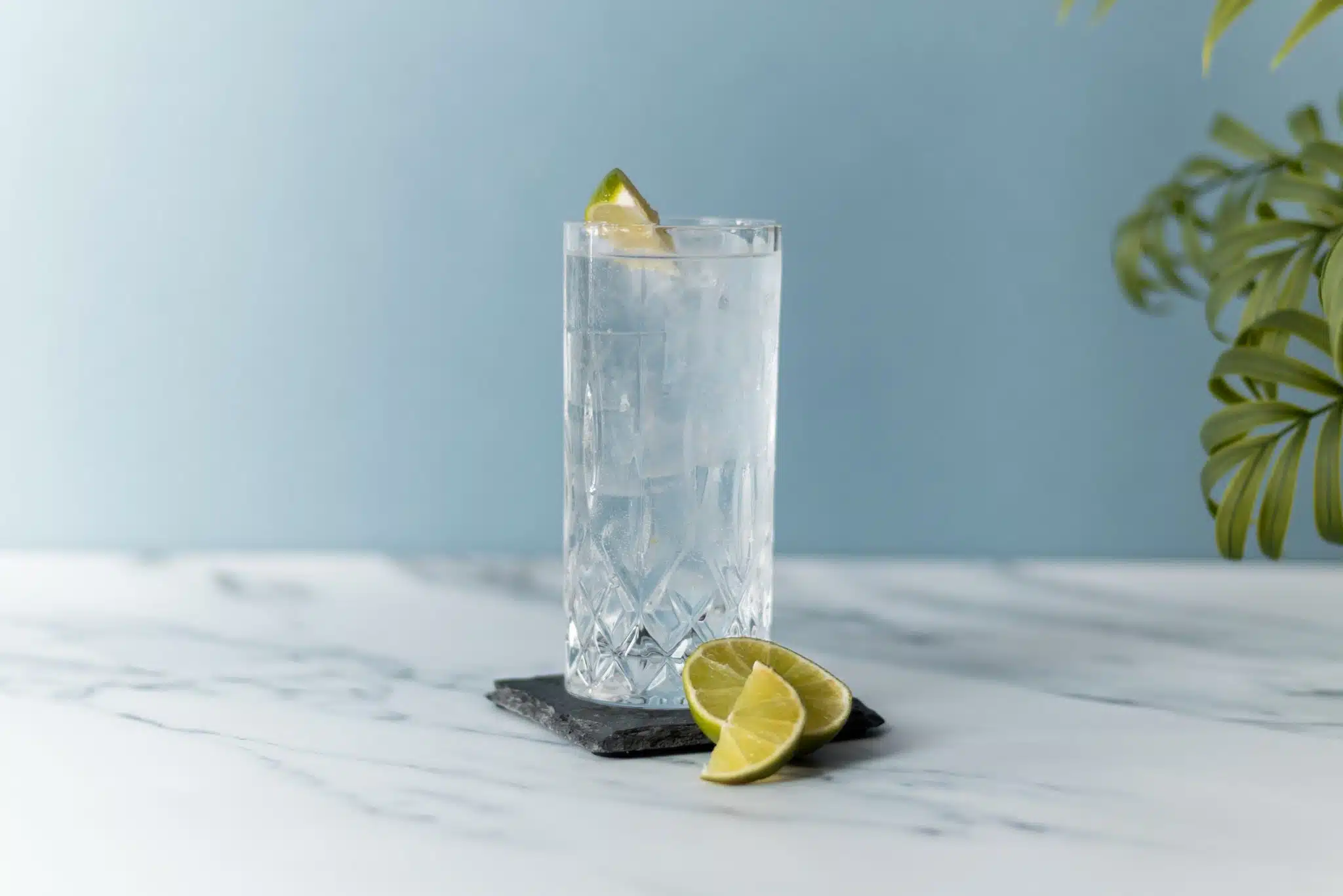 A side shot of a Gin Tonic cocktail in a highball glass on a black stone coaster with lime slices on the side placed on a white marmol table with a light blue background.