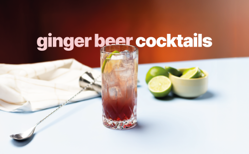 A shot of a ginger beer cocktail in a highball glass on a light blue surface surrounded by limes, a bar spoon, a jigger and a white cloth, in front of a red background