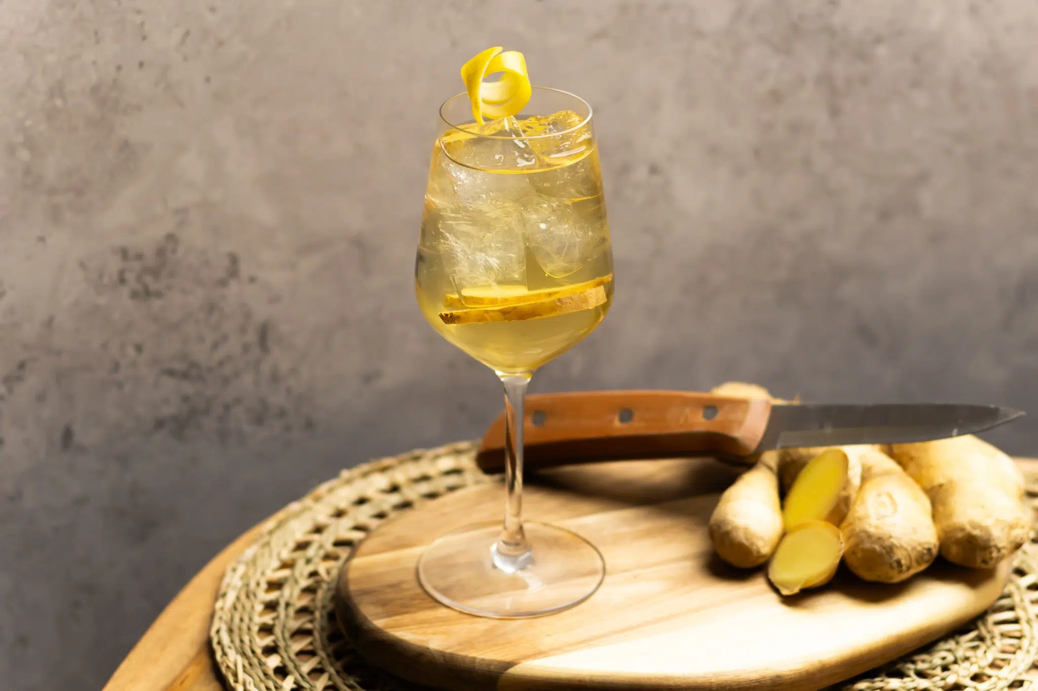 A light yellow beverage with ginger slivers and ice is in a white wine glass on a round wooden cutting board.