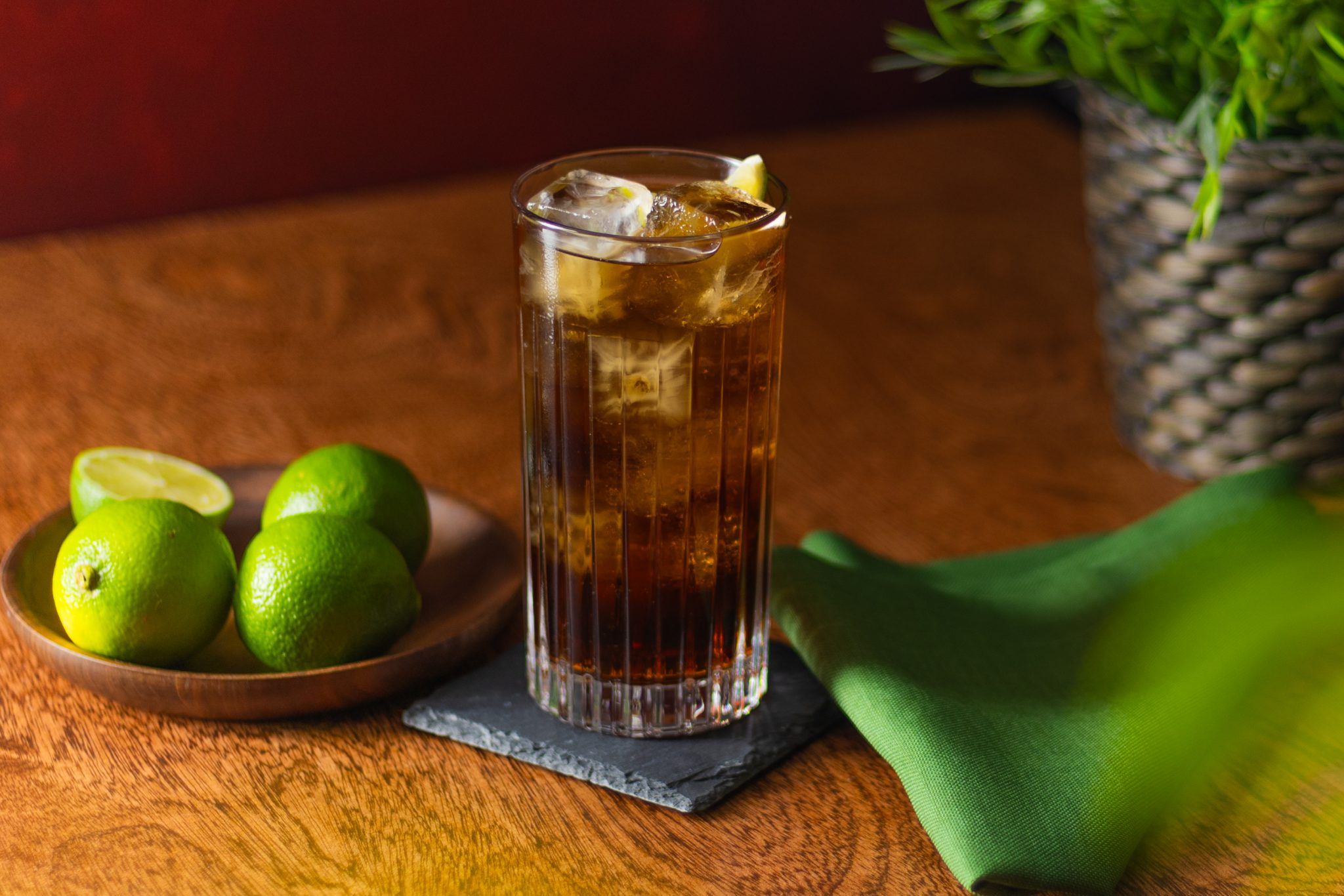 A side shot of a Gold Rum and Coke cocktail in a highball glass on a stone coaster placed on a wooden table with a plate with four limes, a green cloth and a plant around, in front of a red wall.