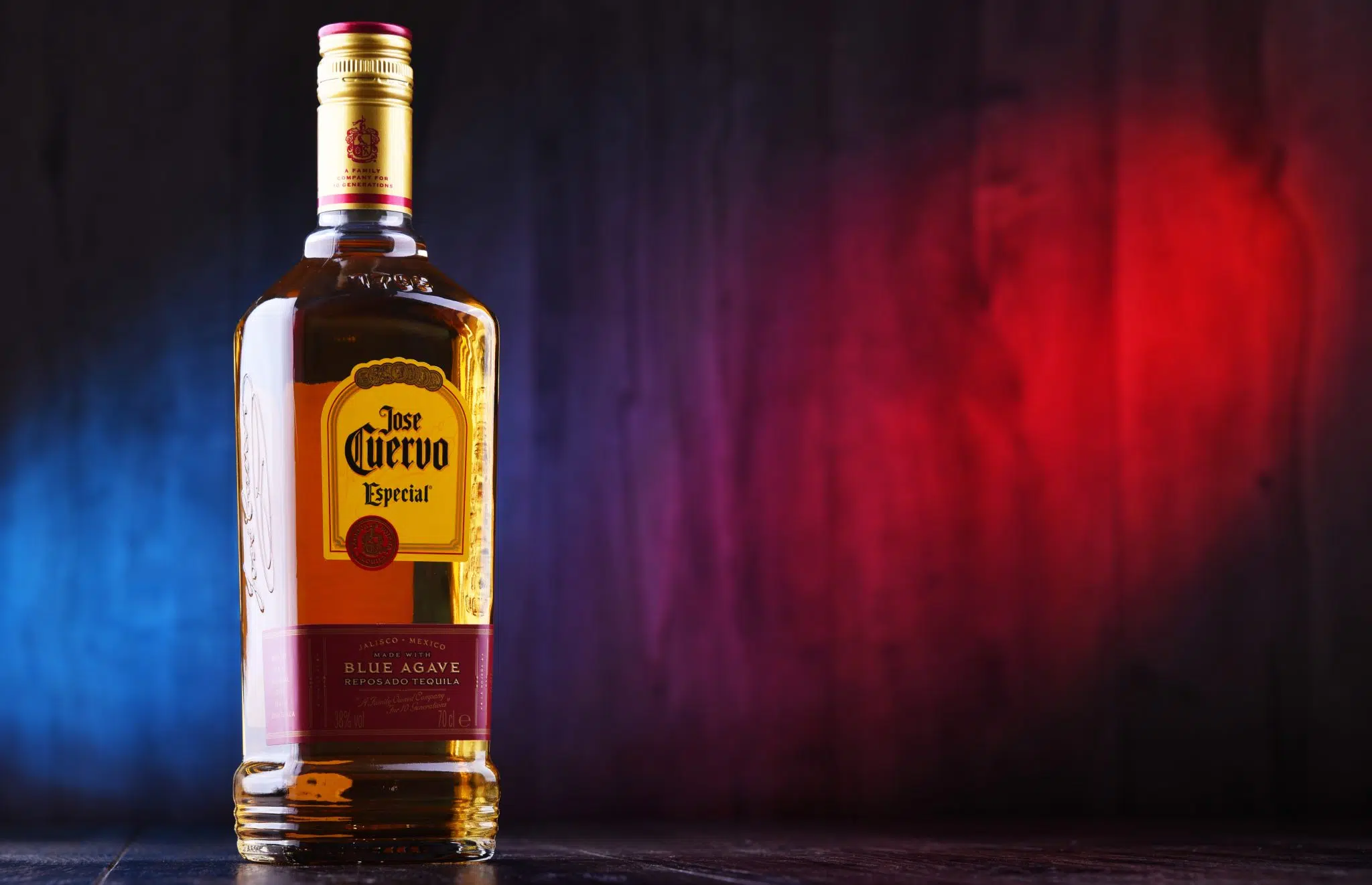 A bottle of a Gold Tequila Jose Cuervo Especial on a shiny dark surface in front of a blue and red background