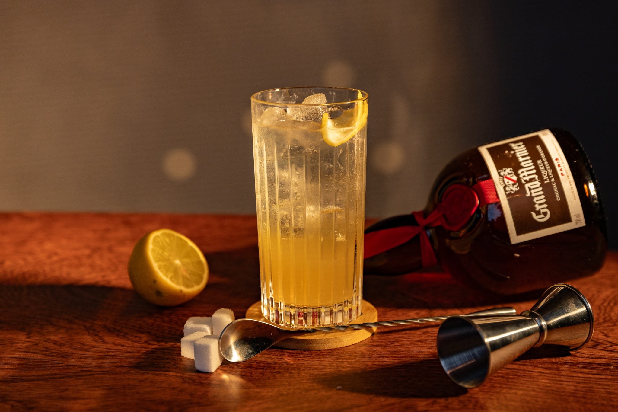 A side shot of a Grand Collins cocktail in a highball glass on a wooden coaster placed on a wooden surface surrounded by a jigger, a bar spoon, five surgar cubes, half a lemon,, and a Grand Marnier bottle
