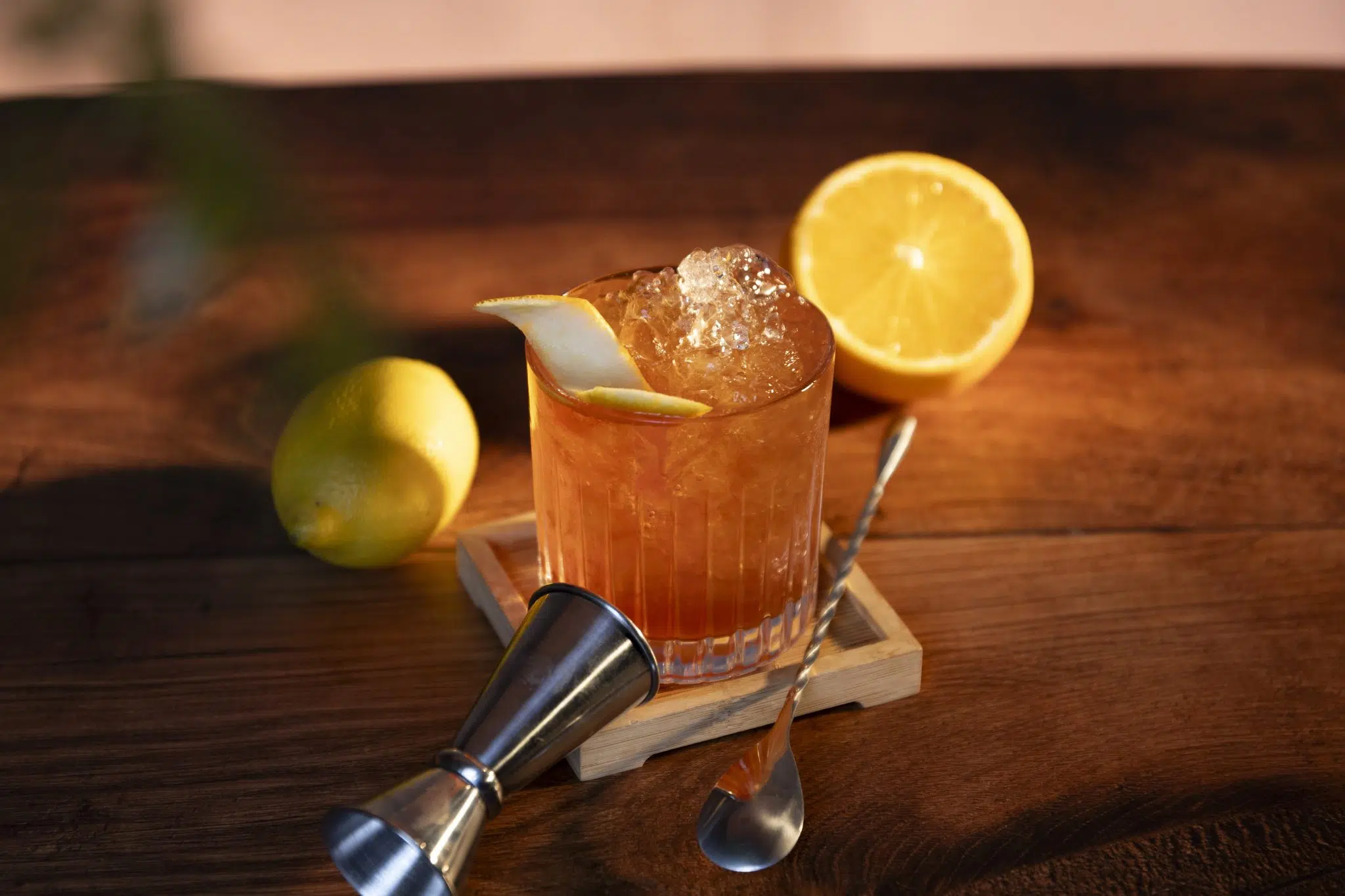 A side shot of a Grand Slam cocktail in an old fashioned glass on a wooden coaster and table surrounded by a lemon, an orange, a jigger and a bar spoon