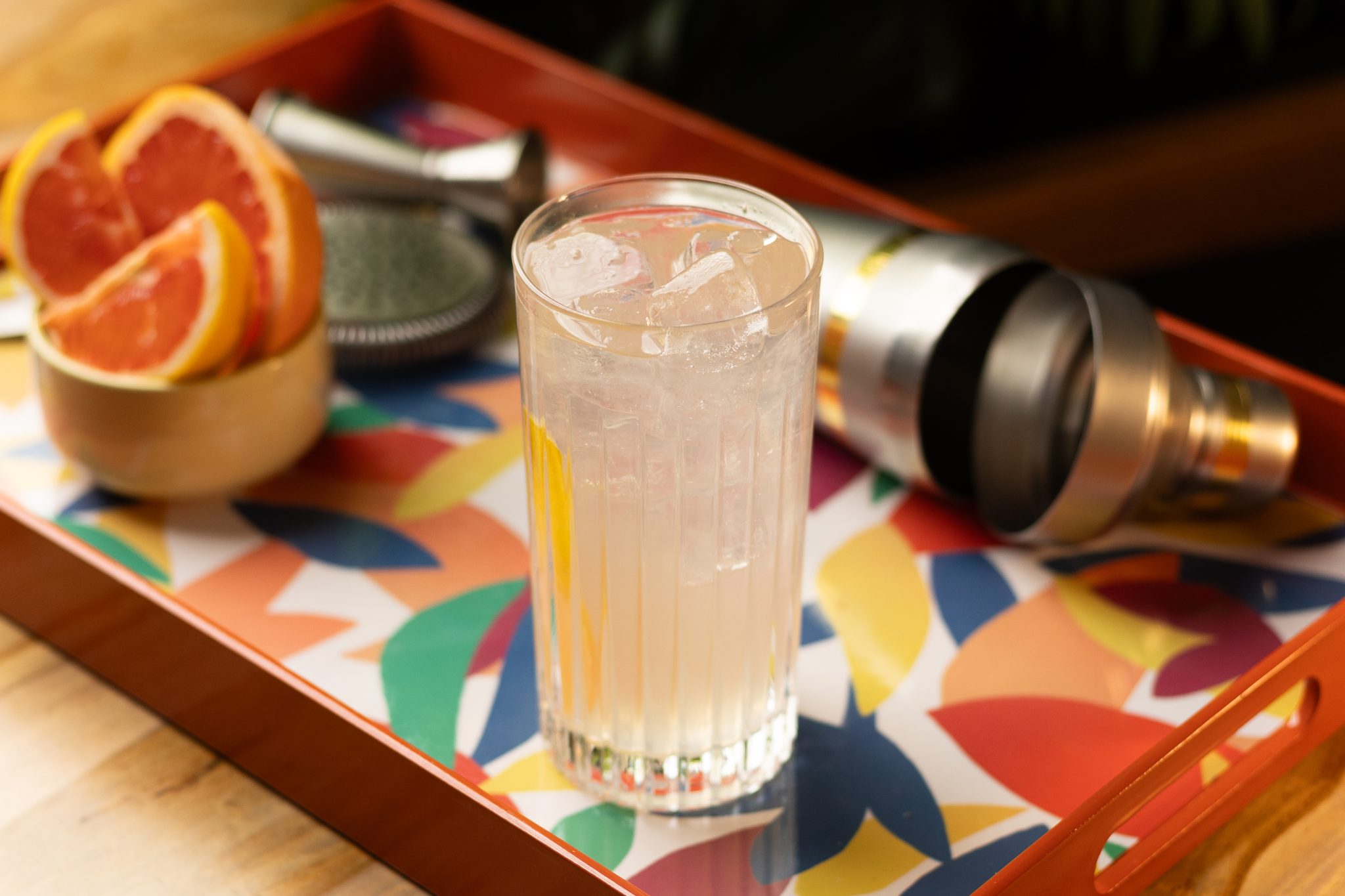 A side shot of a Grapefruit Collins cocktail in a highball glass on a multicolor tray surrounded by a jigger, a shaker, and a bowl with grapefruit wedges.