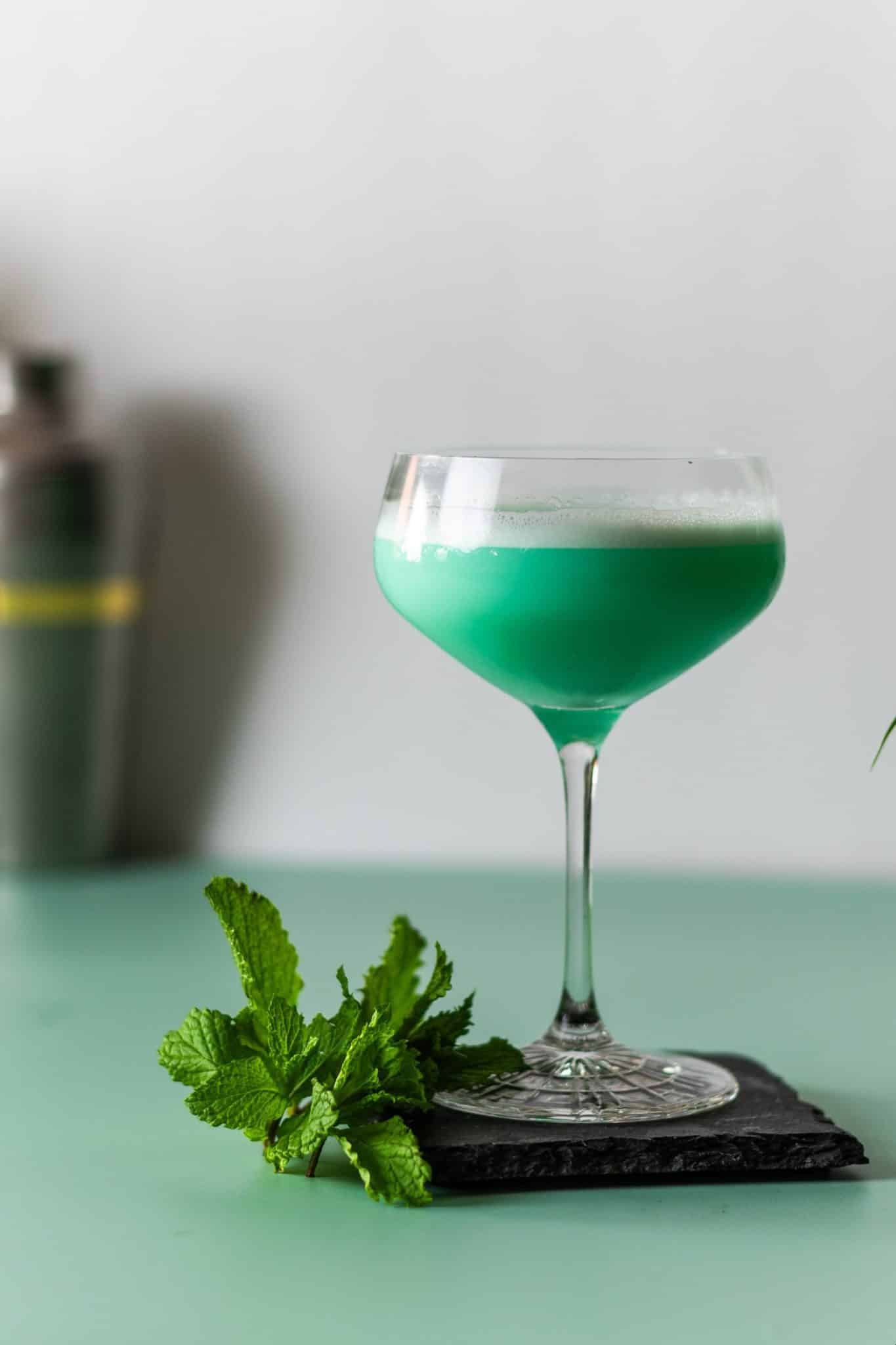 A side shot of a Grasshopper cocktail in a martini glass with a mint sprig on a black stone plate placed on a green table with a shaker on the background.
