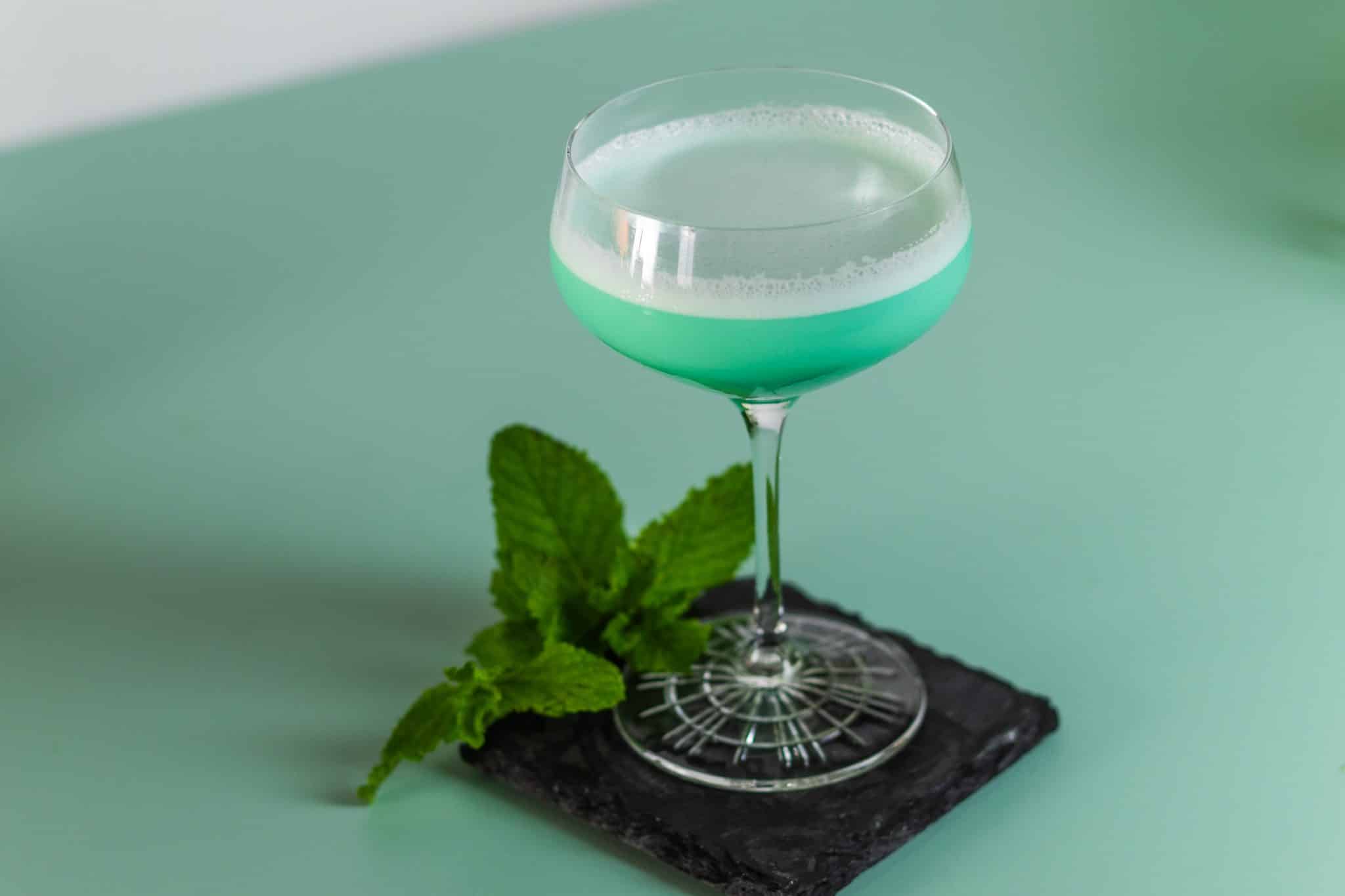 A Grasshopper cocktail, shot from a side, in a martini glass with a mint sprig on a black stone plate placed on a green table and a white wall on the background.
