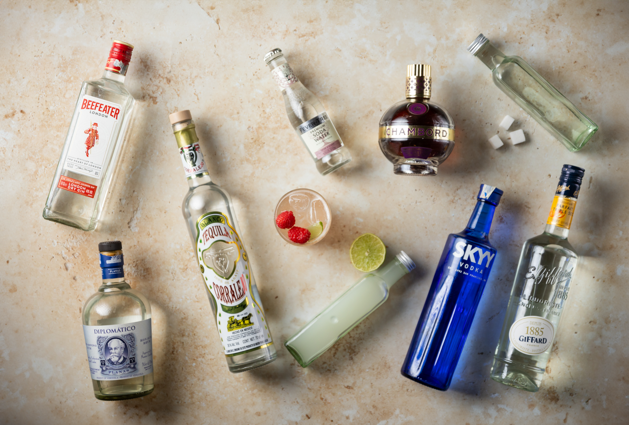 A Grateful Dead drink, shot from above, in a highball glass on a beige surface surrounded by vodka, gin, light rum, tequila, triple sec, simple syrup, raspberry liqueur, lime juice, lime, sugar cubes and soda water