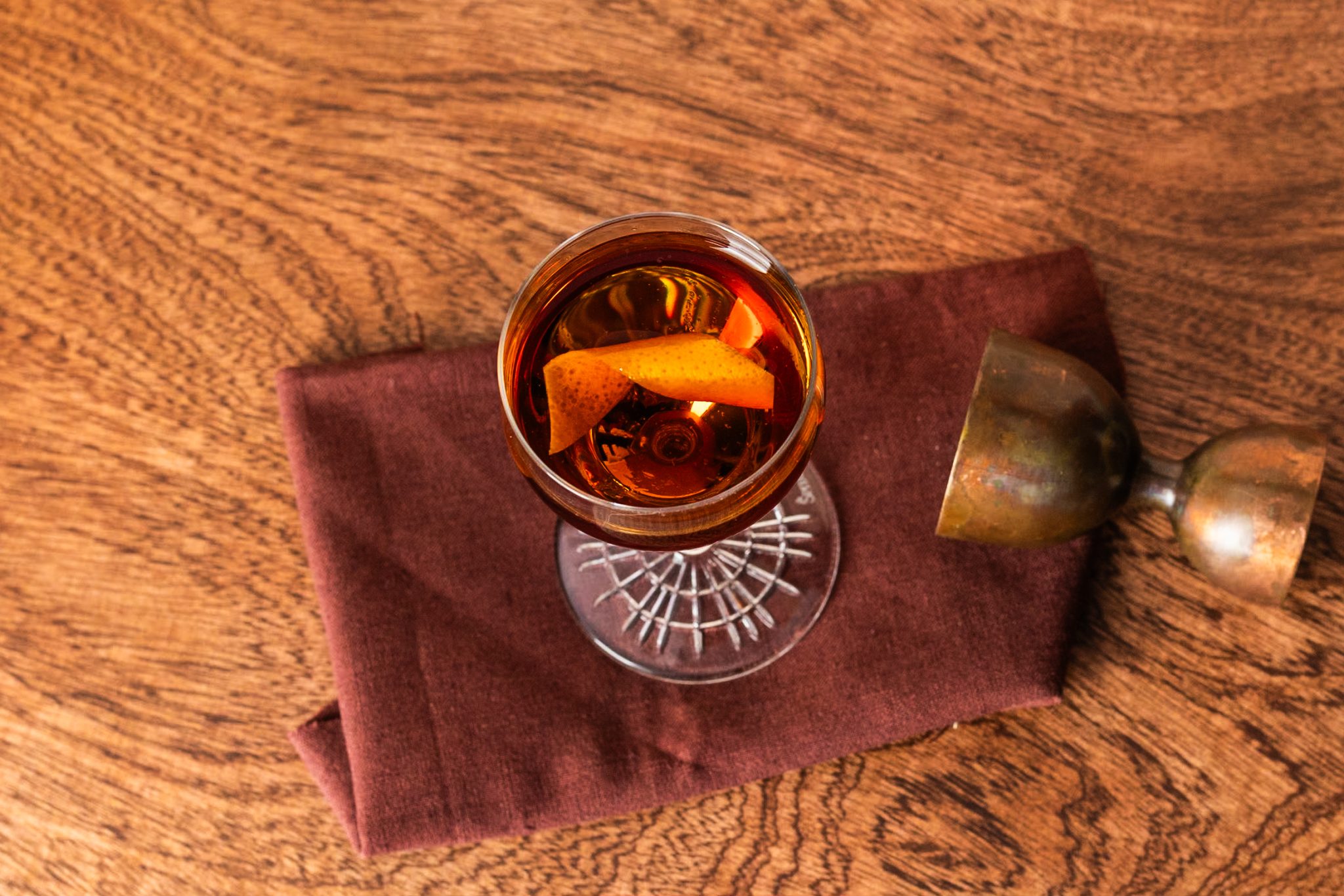 A Hanky Panky cocktail, shot from above, in a coupe glass on a maroon cloth placed on a brown wooden table with a jigger around.