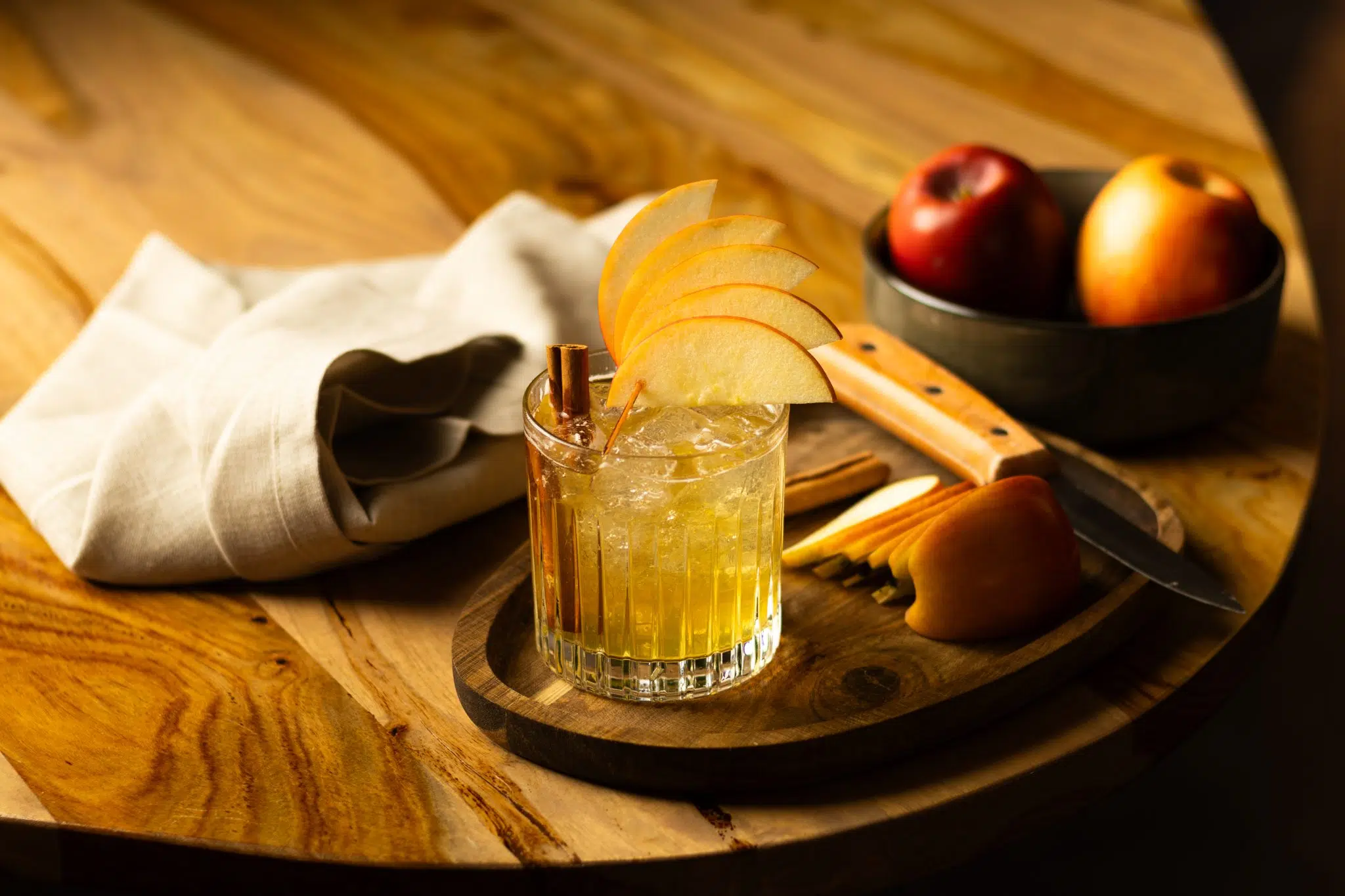 A side shot of a Harvest Punch cocktail in an old fashioned glass on a wooden board placed on a wooden table surrounded by two apples, a white cloth and apple slices.