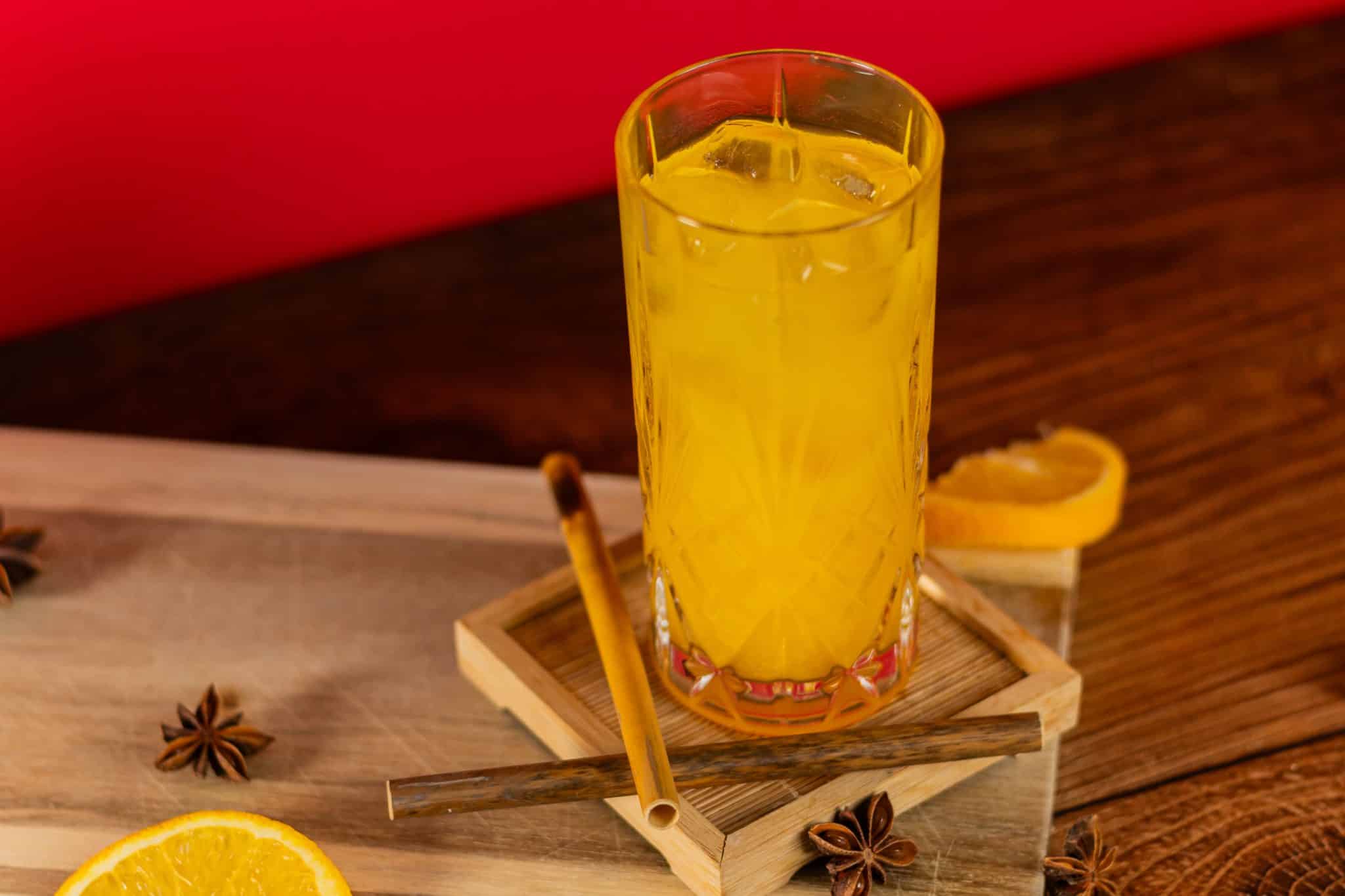 A side shot of a Harvey Wallbanger cocktail in a highball glass with two straws on a wooden coaster placed on a wooden table and orange slices around.