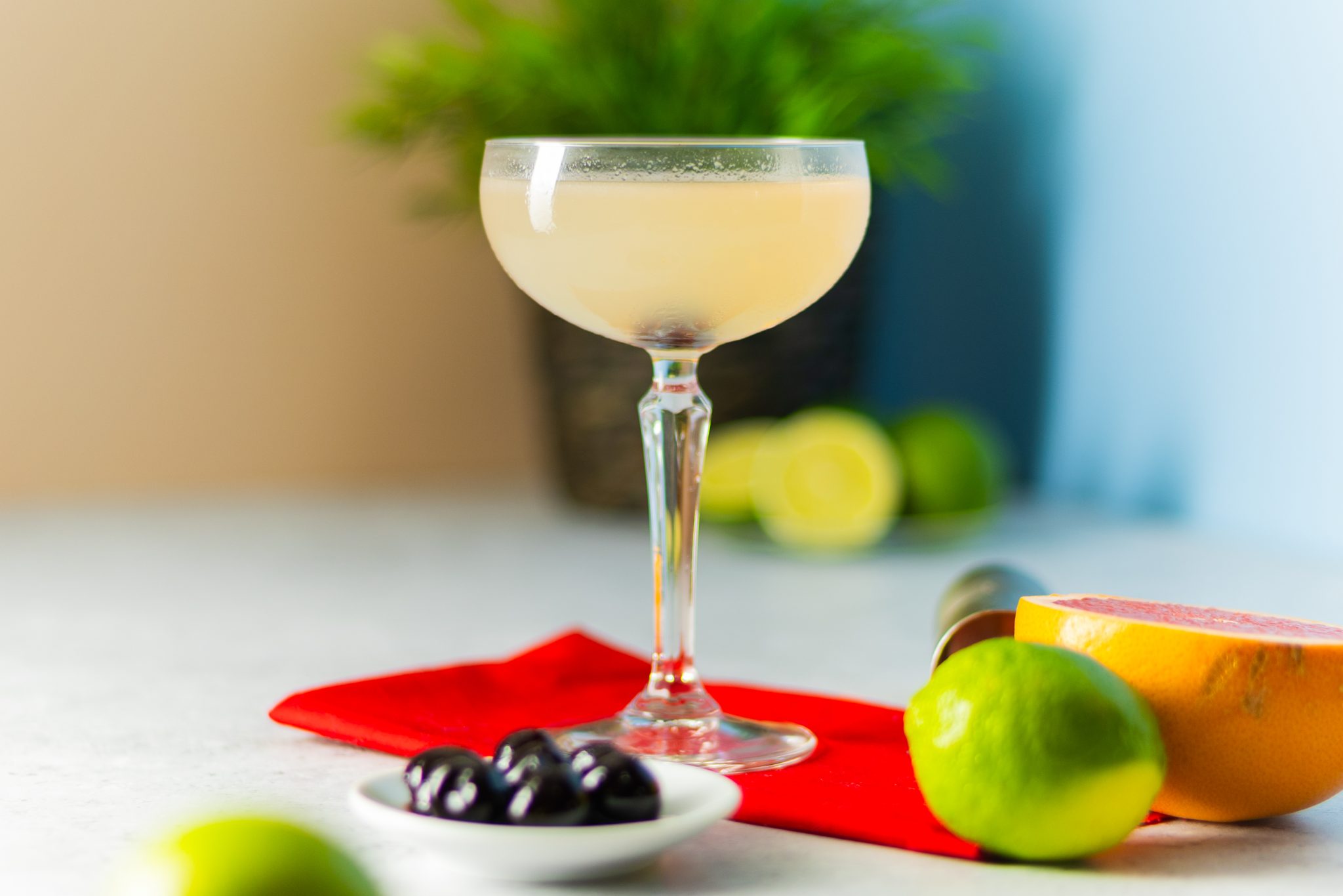 A side shot of a Hemingway Daiquiri cocktail in a cocktail glass on a red cloth placed on a white table with a plate full with maraschino cherries, two limes, and half grapefruit around, in front of a yellow and blue wall.