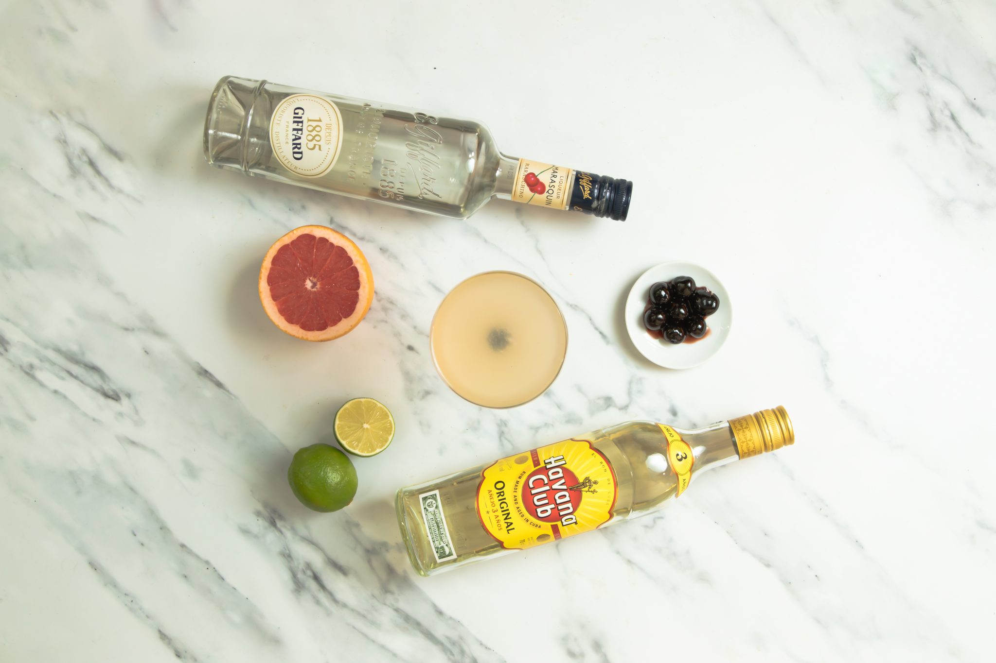 A Hemingway Daiquiri cocktail, shot from above, in a cocktail glass on a white marmol table surrounded by a Maraschino Liqueur bottle, a White Rum bottle, half grapefruit, two limes, and a plate full with Maraschino cherries.