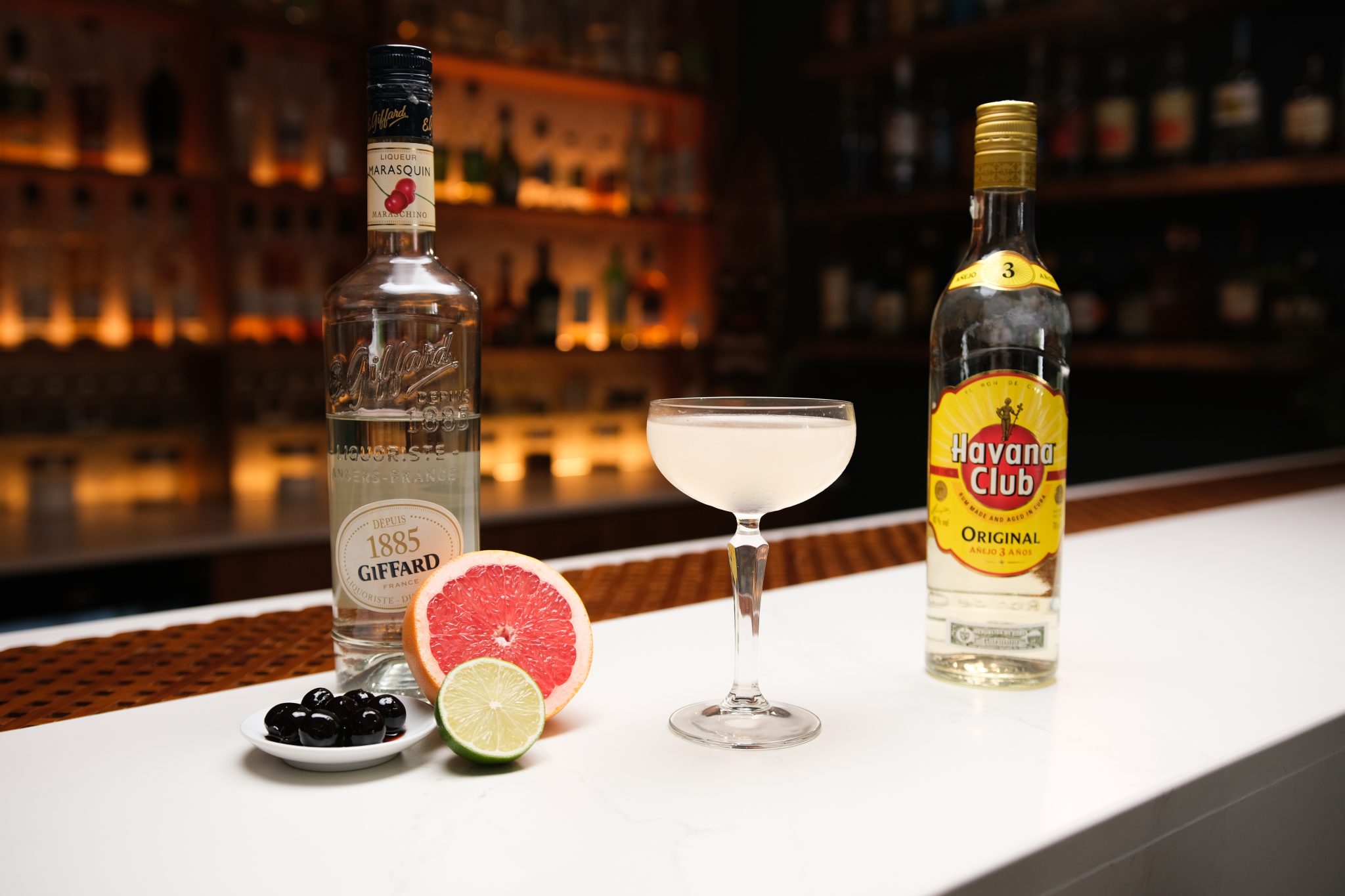 White Rum, Maraschino Liqueur, half lime, half grapefruit, and a plate full with Maraschino cherries laid out on a white bar table.