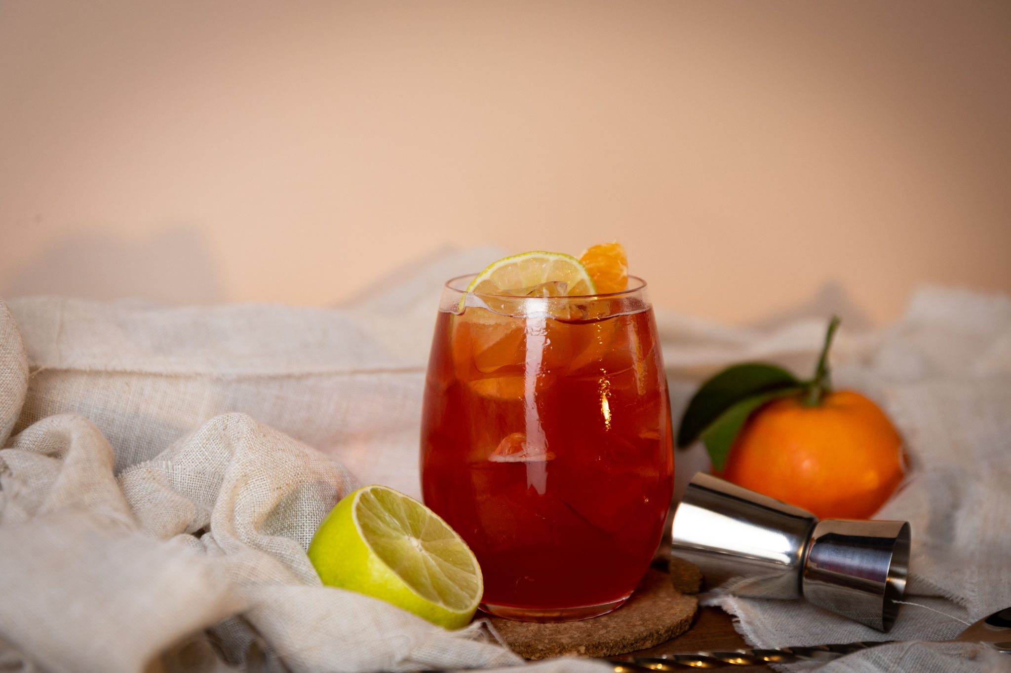 A side shot of a Holiday Margarita cocktail in a rocks glass on a cork coaster surrounded by a jigger, an orange, a white cloth and half a lemon